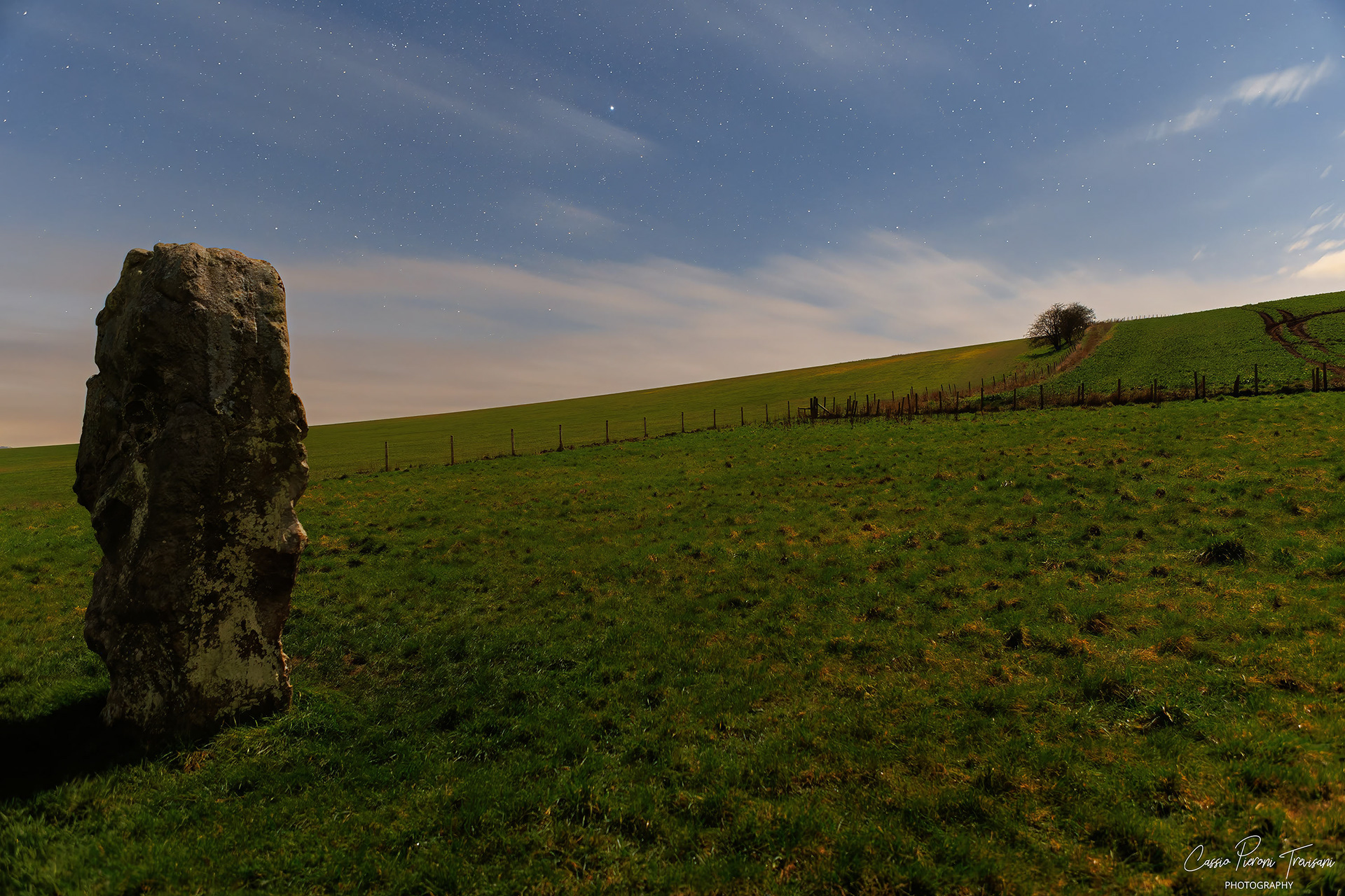 A single megalith stands prominently in the foreground, silhouetted against the sloping Wiltshire hills and a sky brushed with faint starlight.