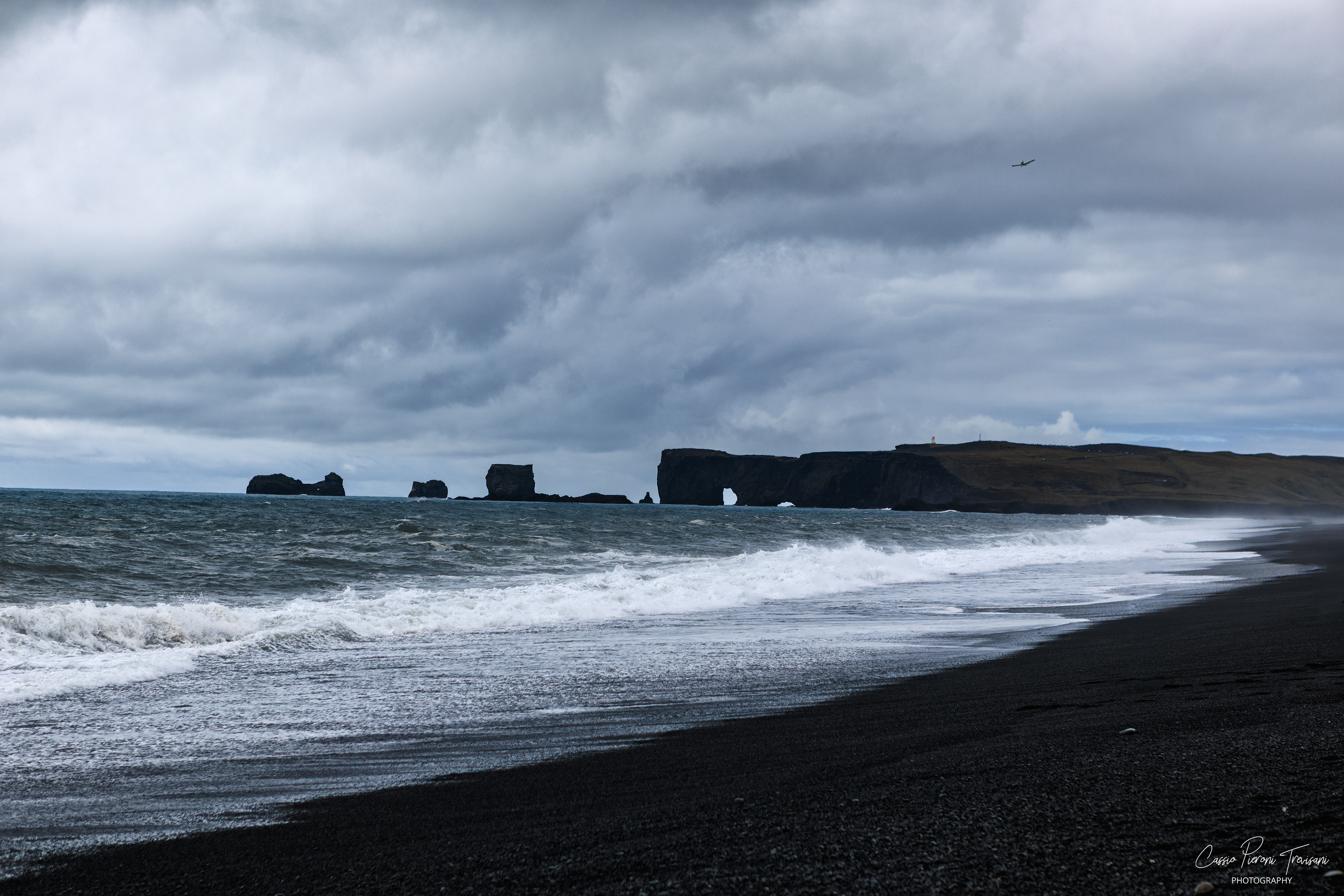 Waves roll across the black sand of Reynisfjara beneath a dramatic sky, with distant sea stacks on the horizon.
