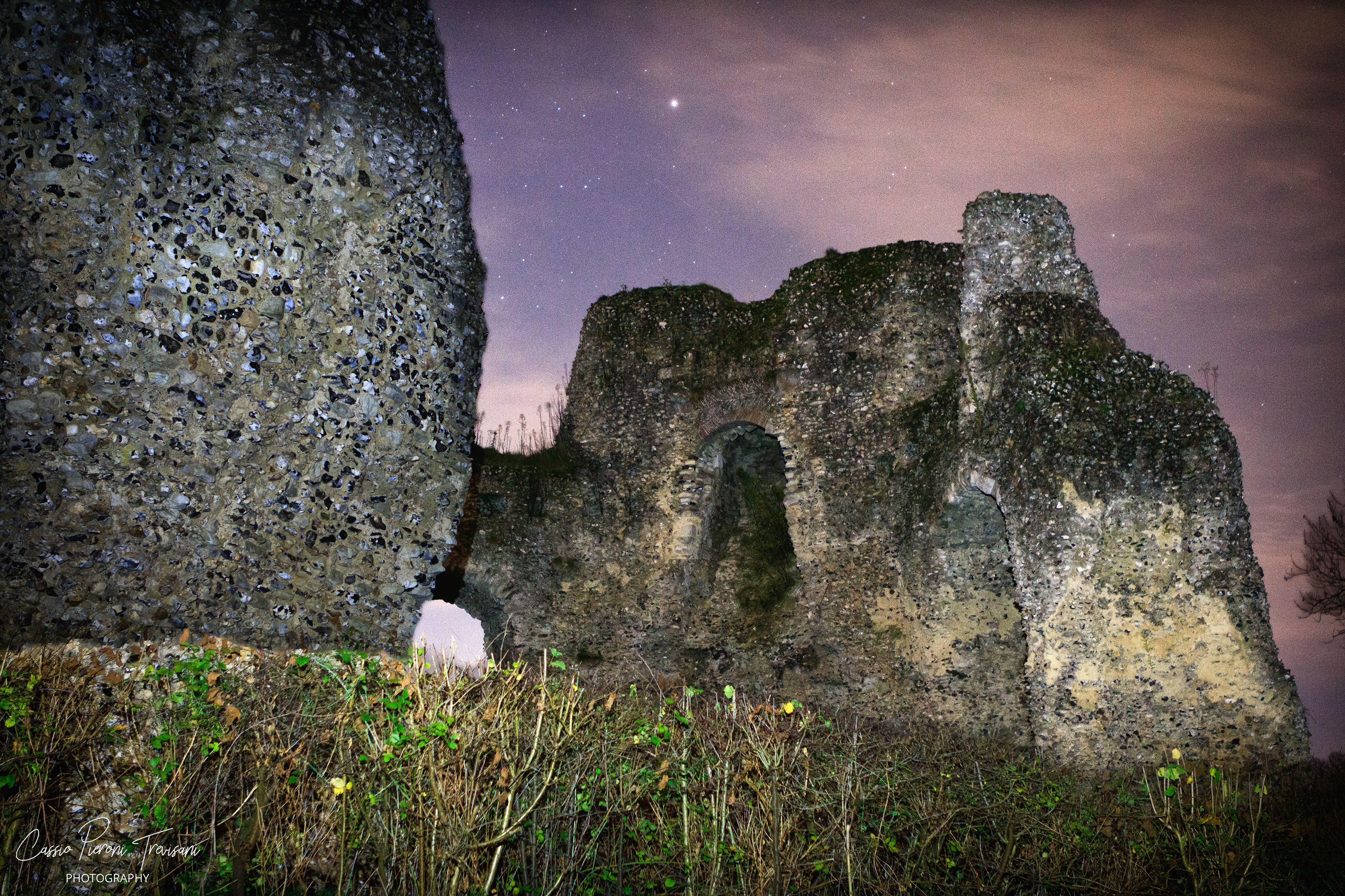 Close nighttime view of Odiham Castle’s crumbling stone walls with archways beneath a pink-tinted sky.