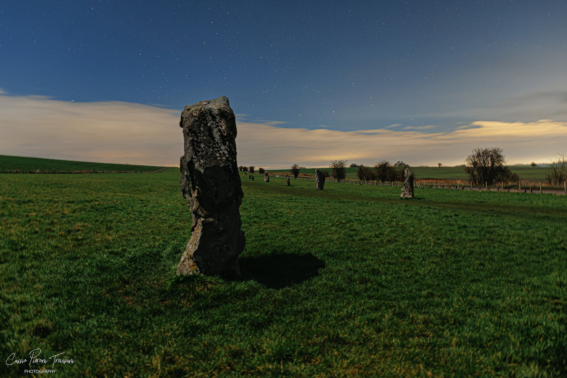 A leading stone anchors the frame as West Kennet Avenue stretches into the distance beneath a star-filled sky, illuminated by soft horizon glow and drifting clouds.
