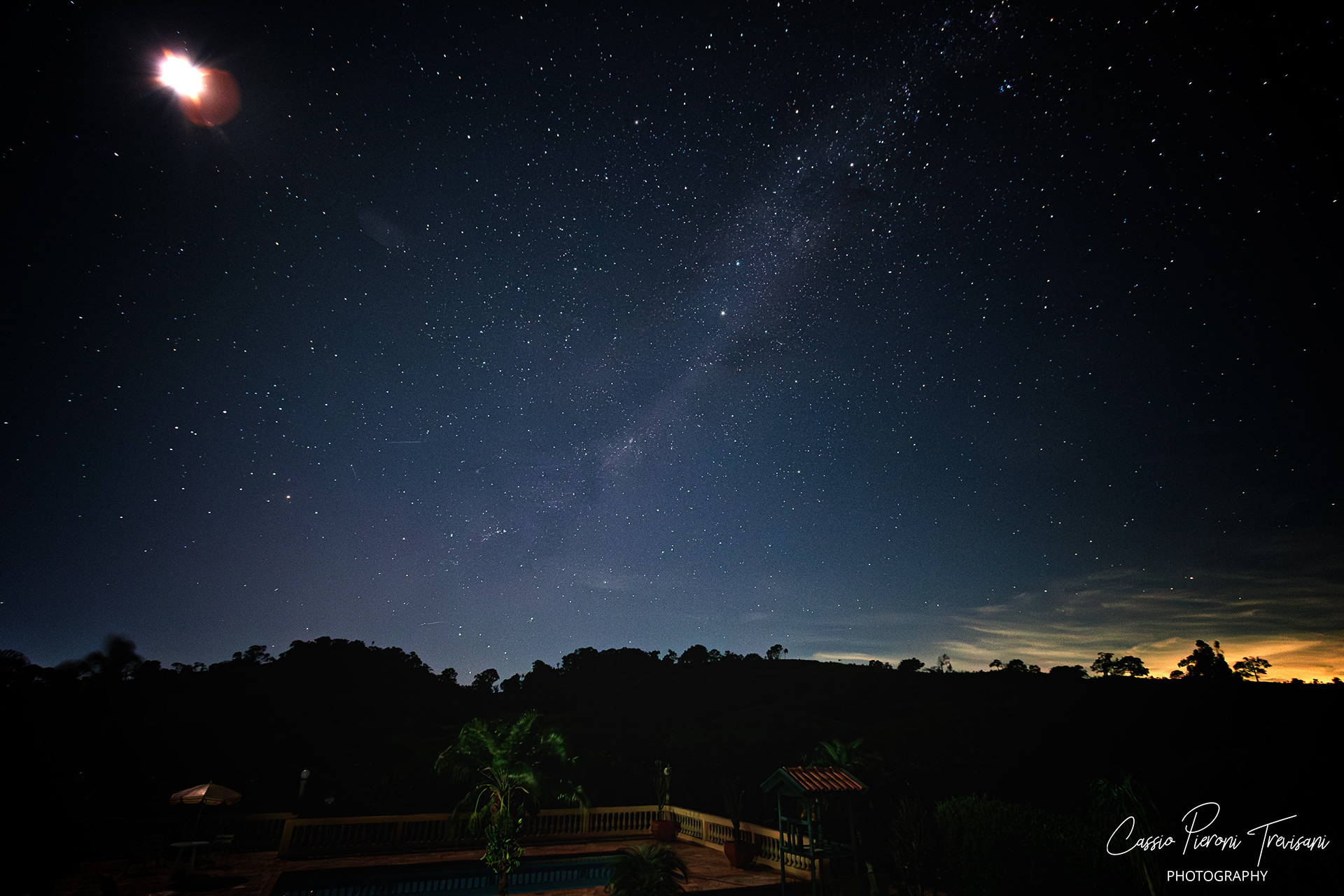 A star-filled night sky arches over a rural landscape with silhouetted trees and a softly lit farm, as the Milky Way fades gently across the heavens in Minas Gerais.