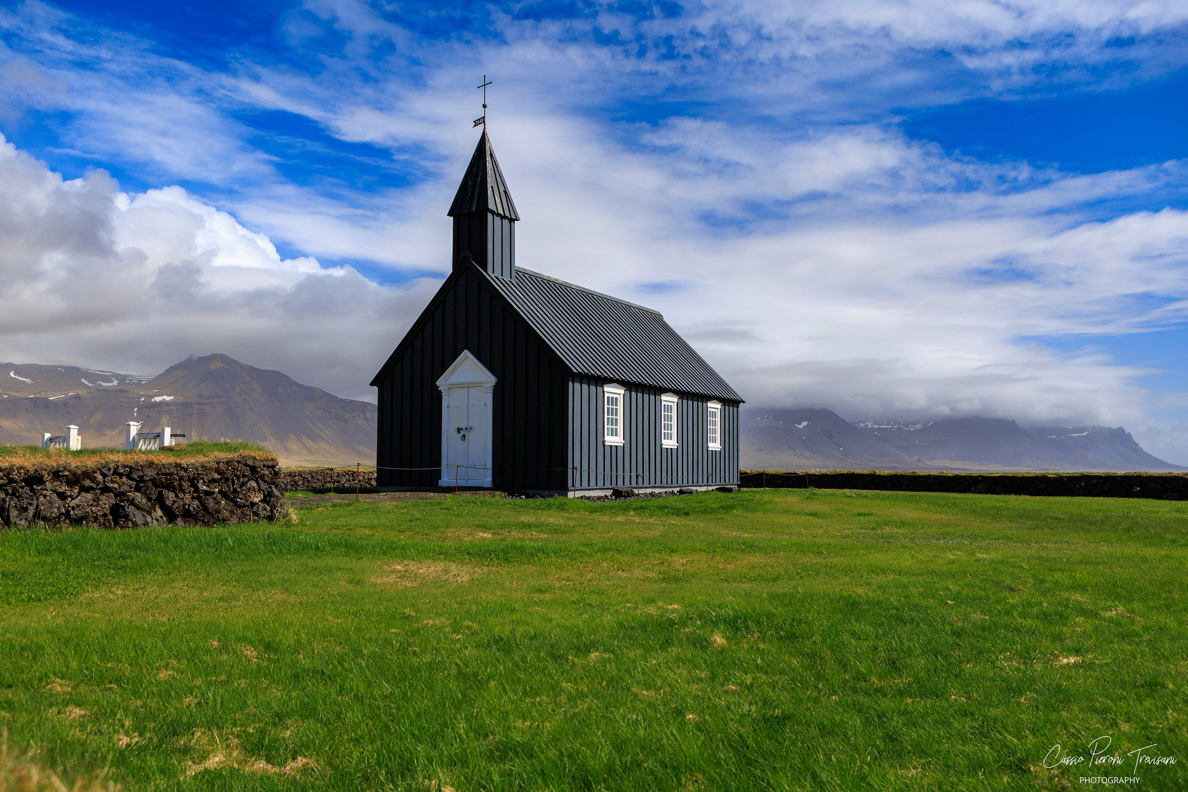 A solitary black church (Búðakirkja) stands against sweeping clouds and distant mountains, capturing the quiet elegance of Iceland’s rural landscape.