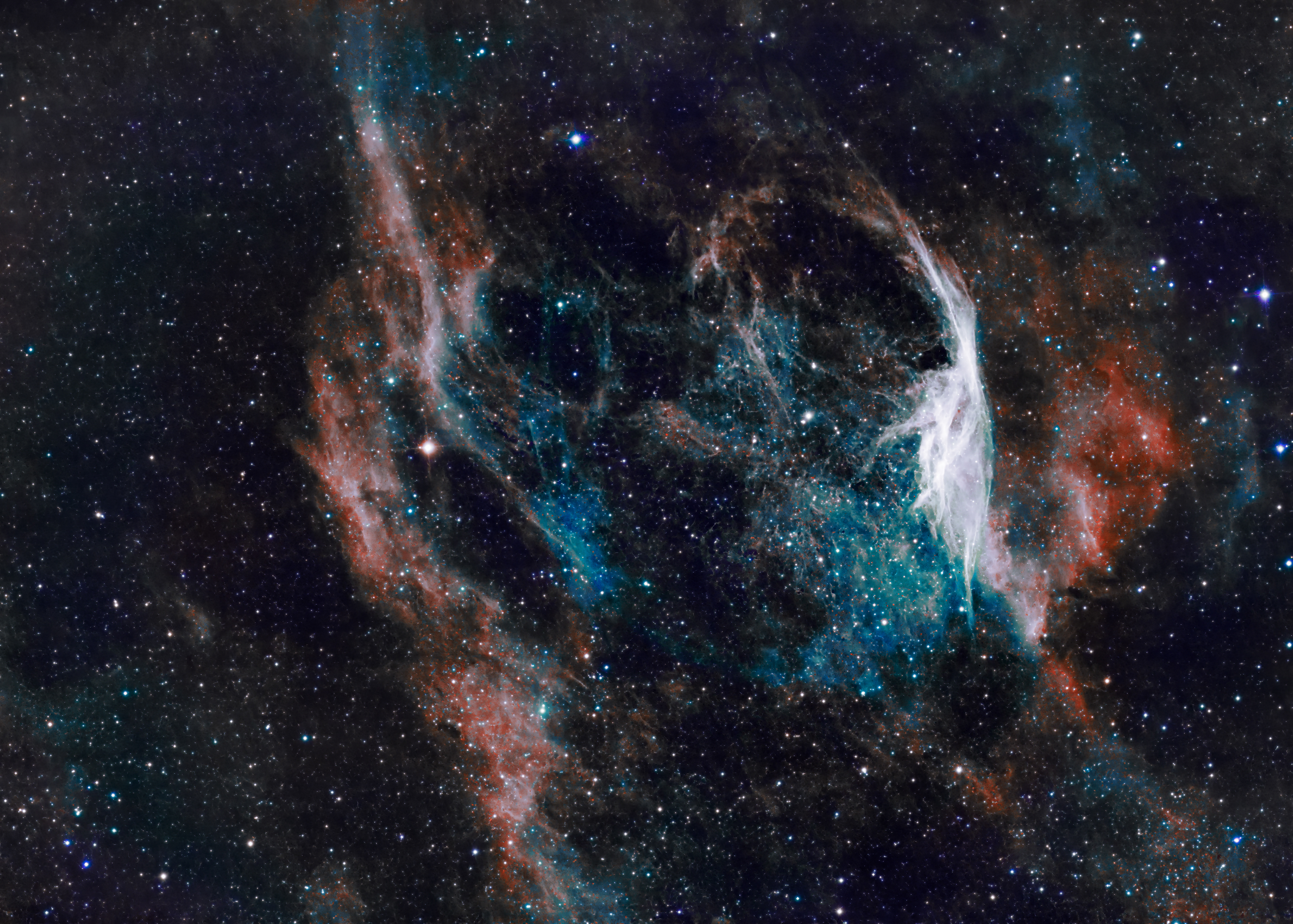 Astrophotograph of RCW 104, showing a delicate bubble-shaped nebula with intricate filaments. The structure glows in shades of blue and cyan in the interior, surrounded by warmer orange and reddish gas, all set against a dense background of stars.