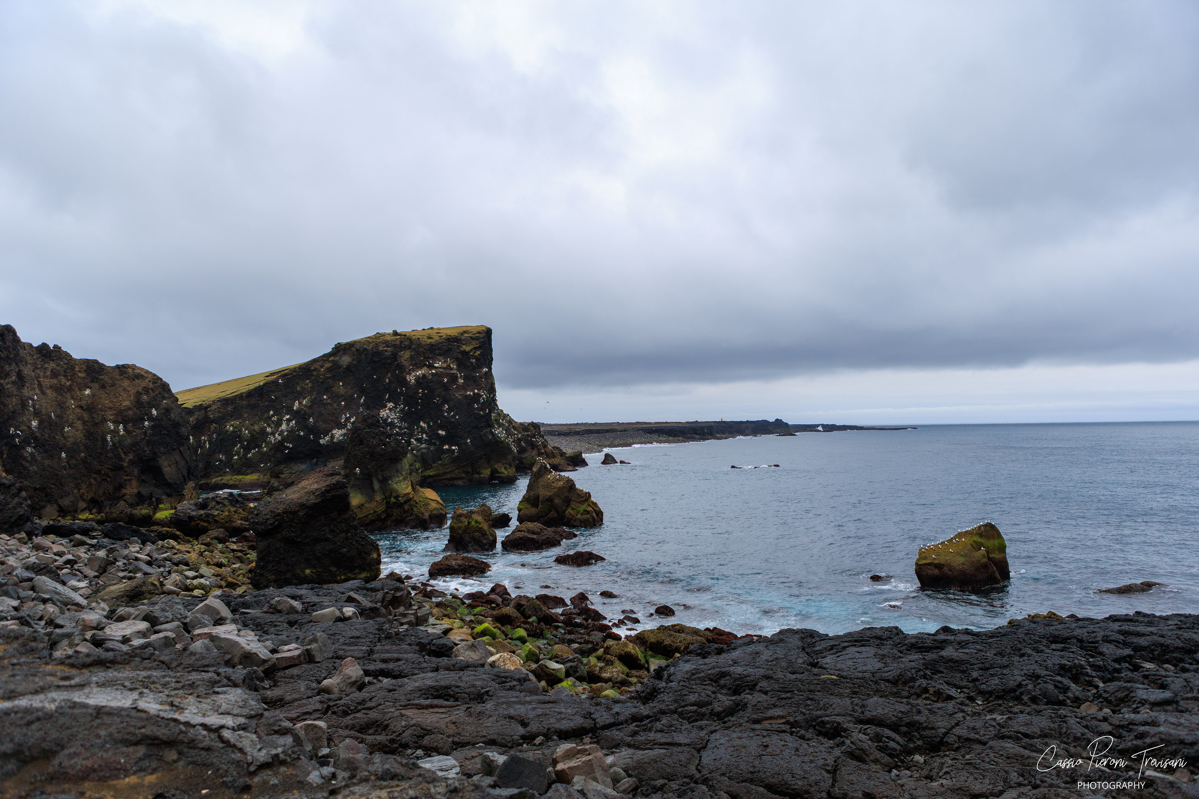 The rugged coastline of Valahnúkamöl stretches into the Atlantic, where volcanic cliffs meet calm blue water beneath low clouds.