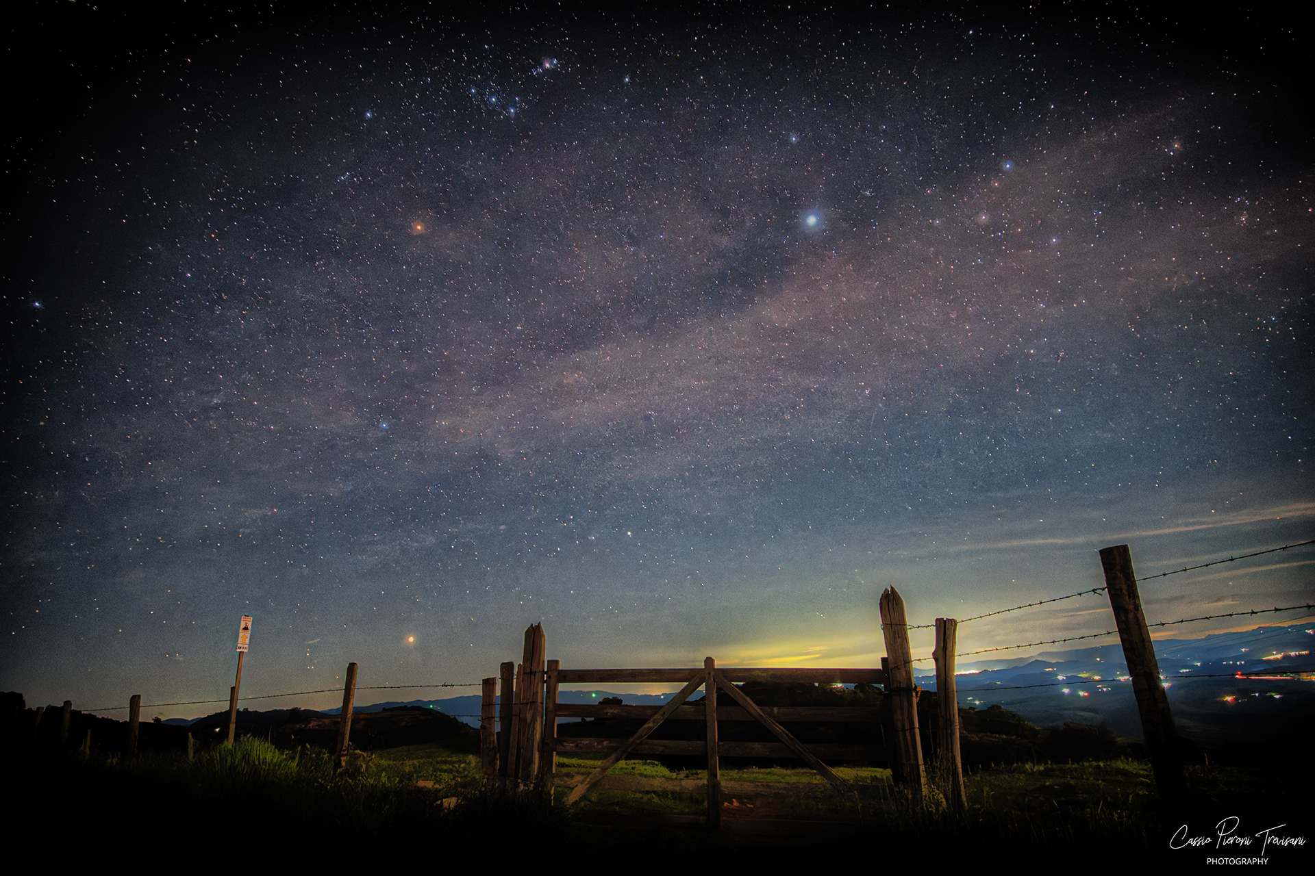 Milky Way arching over a wooden gate in a rural landscape at night, with distant city lights on the horizon in Jacutinga, Minas Gerais, Brazil.