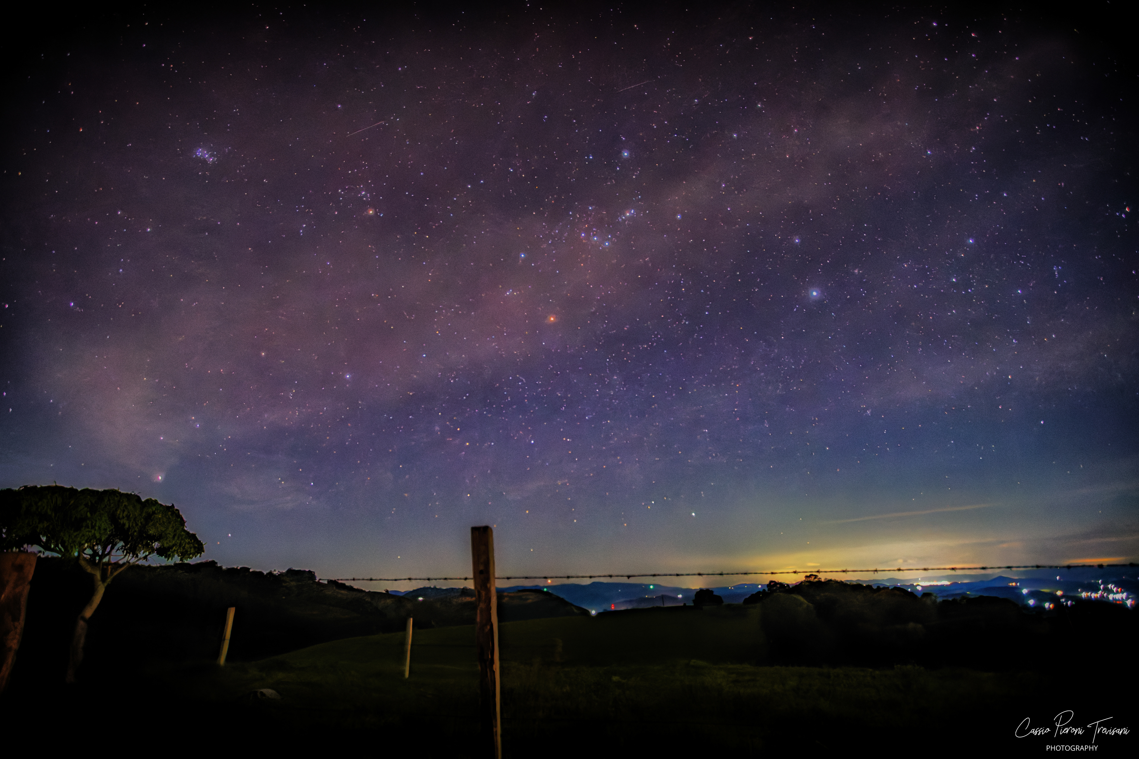 Milky Way stretching across the night sky above rural hills and fence posts, with distant city lights in Jacutinga, Minas Gerais, Brazil.