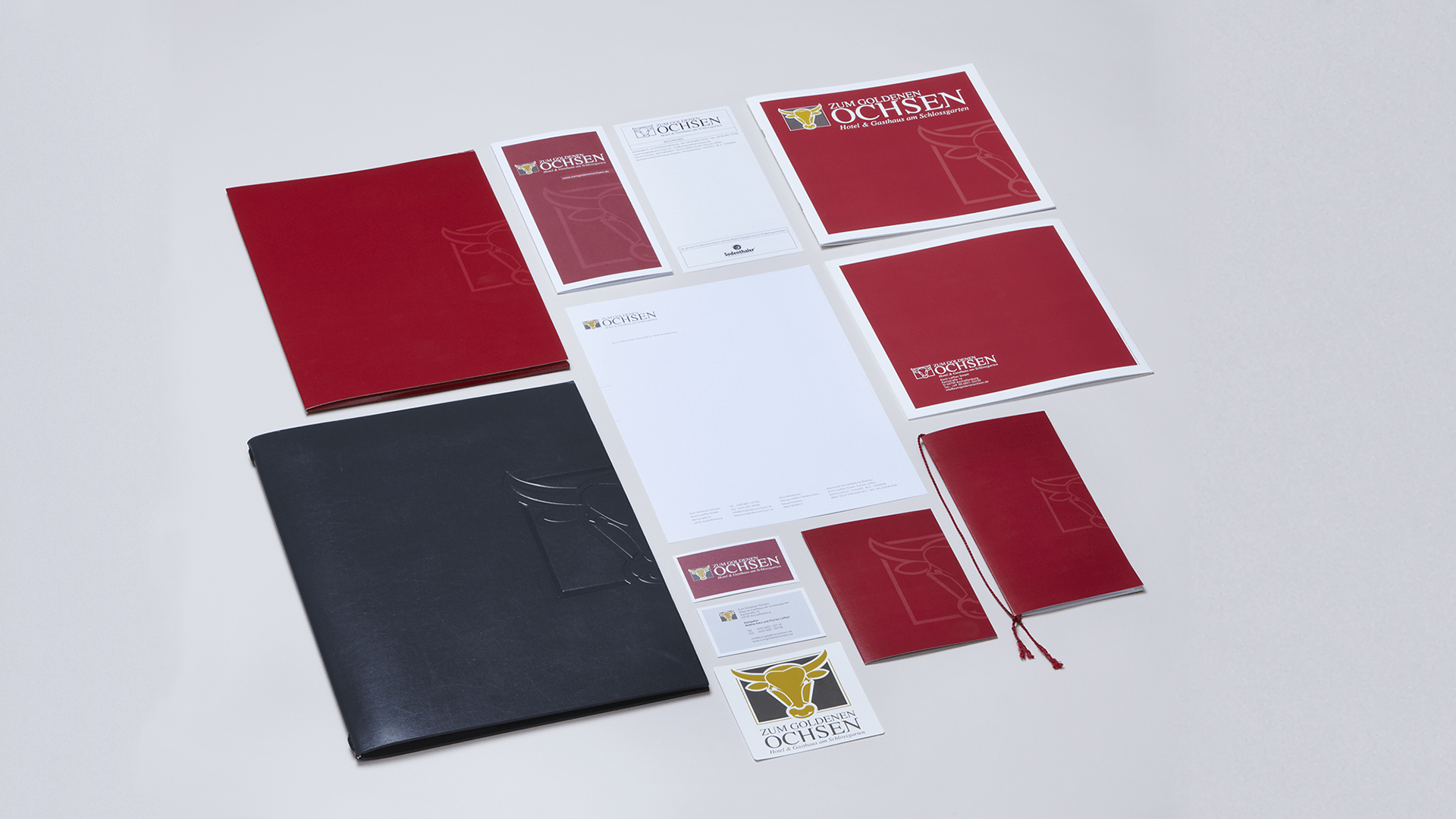 Übersicht der Geschäftsausstattung vom Hotel & Gasthaus Zum Goldenen Ochsen: rote und schwarze Mappen, Briefpapier, Visitenkarten, Notizblöcke und weitere Printmedien im einheitlichen Corporate Design, sauber auf hellem Hintergrund arrangiert.
