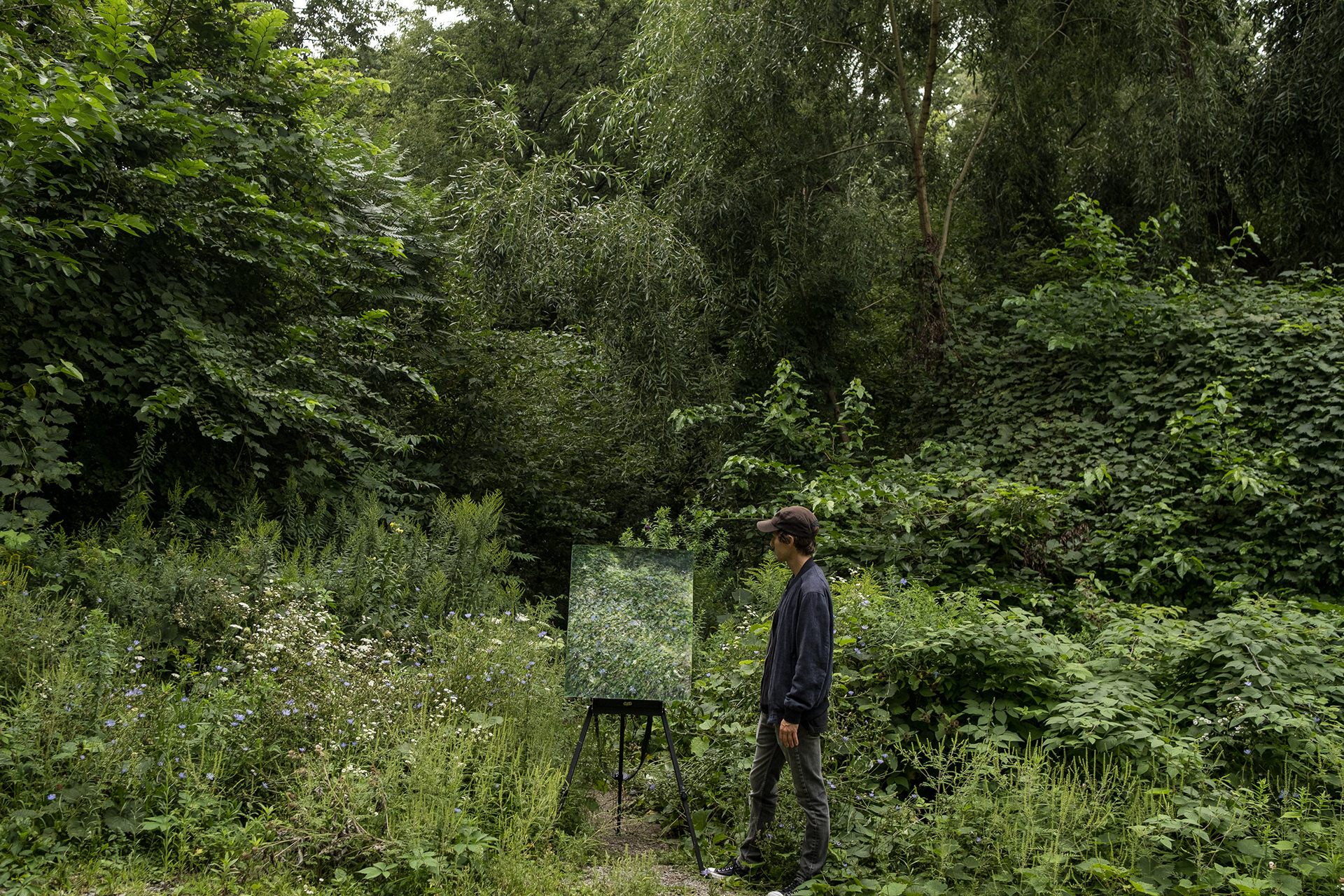 Curt standing in a patch of verdant green wild flowers, behind his easel with a painting of the wildflowers in the summer