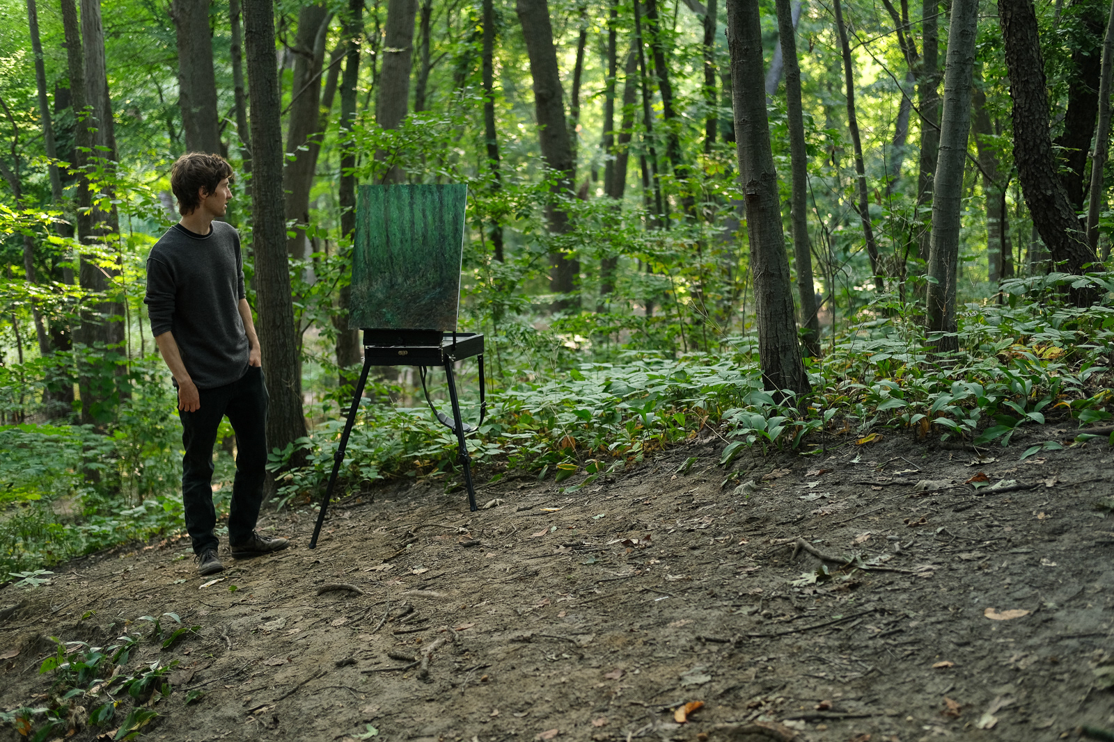 Curt standing in the forest early morning, behind his easel with a painting of the forest in the summer