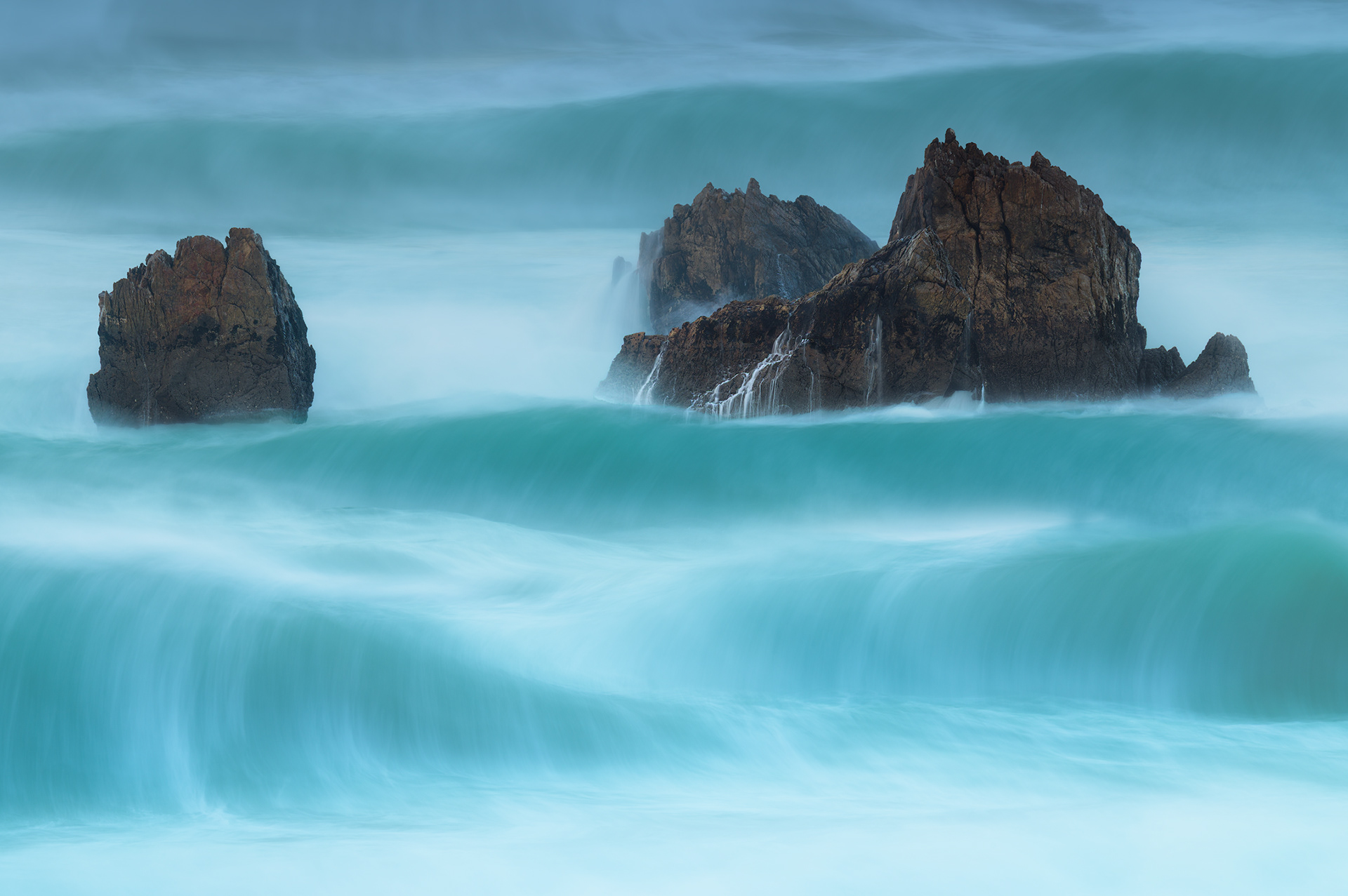 Long exposure seascape photography at Muros del Nalón, Asturias, Spain. High-contrast dark jagged rocks in the Cantabrian Sea surrounded by misty, silky turquoise waves with a motion blur effect. Professional landscape shot during the Goretti storm, featuring emerald water tones, sharp rock textures, and an ethereal atmosphere 
