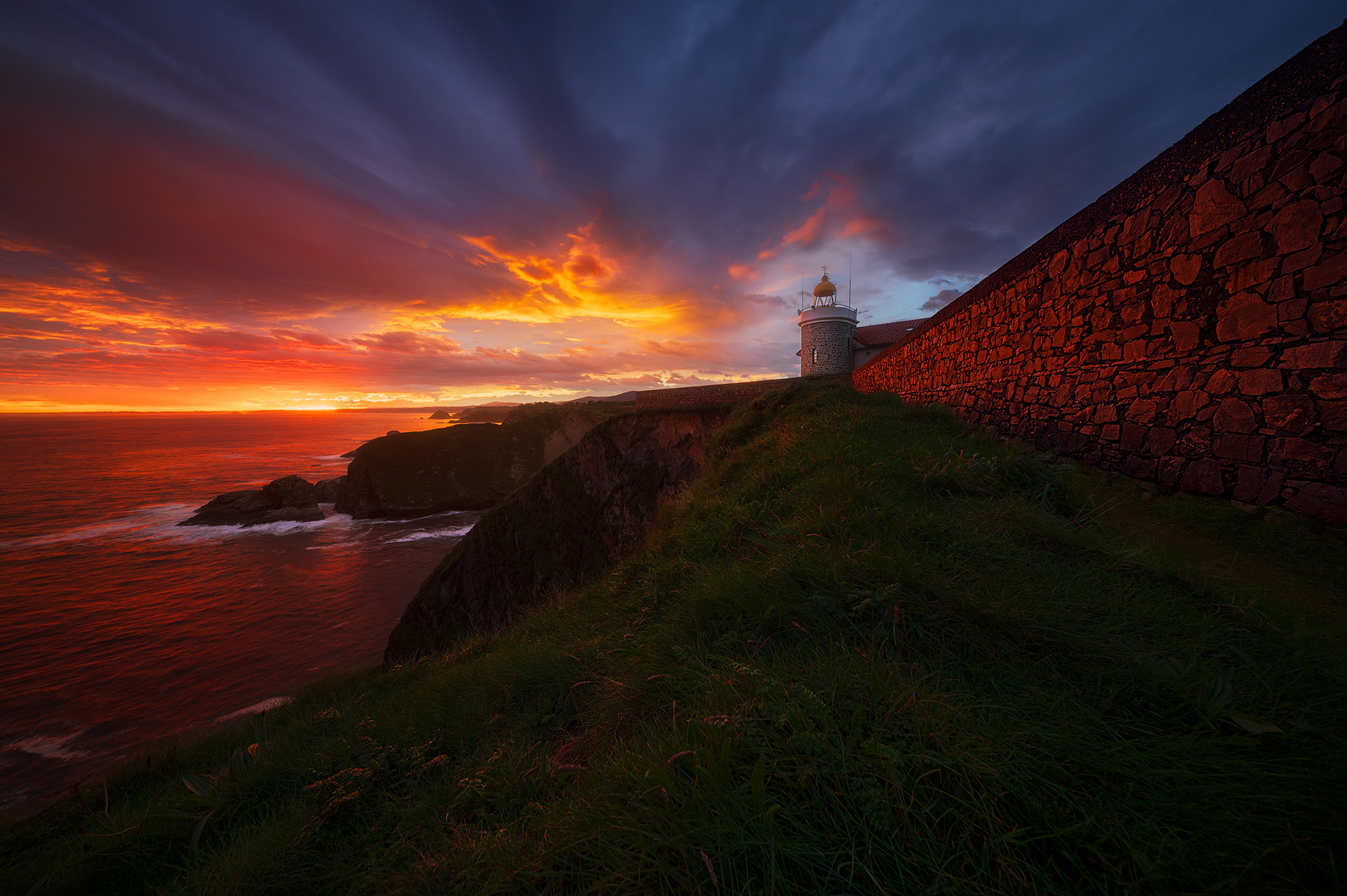 Amanecer en el Faro del Cabo Vidio, con el cielo teñido de rojo y amarillo, reflejando su luz sobre el mar Cantábrico y las rocas costeras