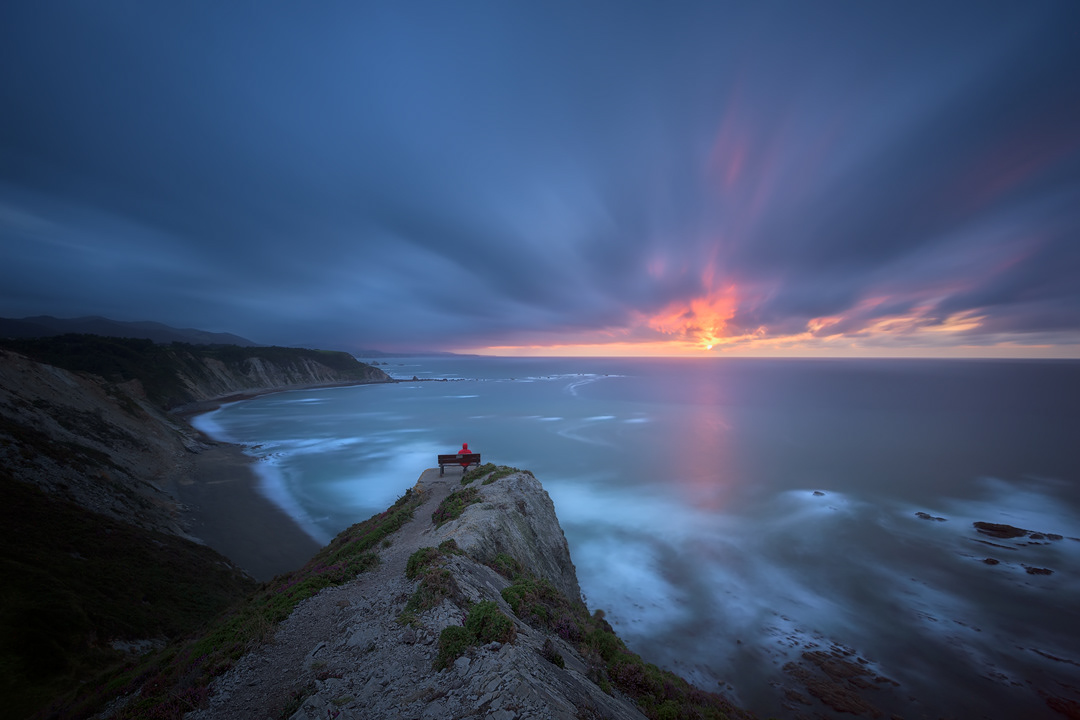 Atardecer de película en el mirador del Sablón, Oviñana, Asturias, con nubes a la fuga, el sol poniéndose y reflejándose en el mar, mientras el cielo se tiñe de tonos naranjas, anaranjados y azules
