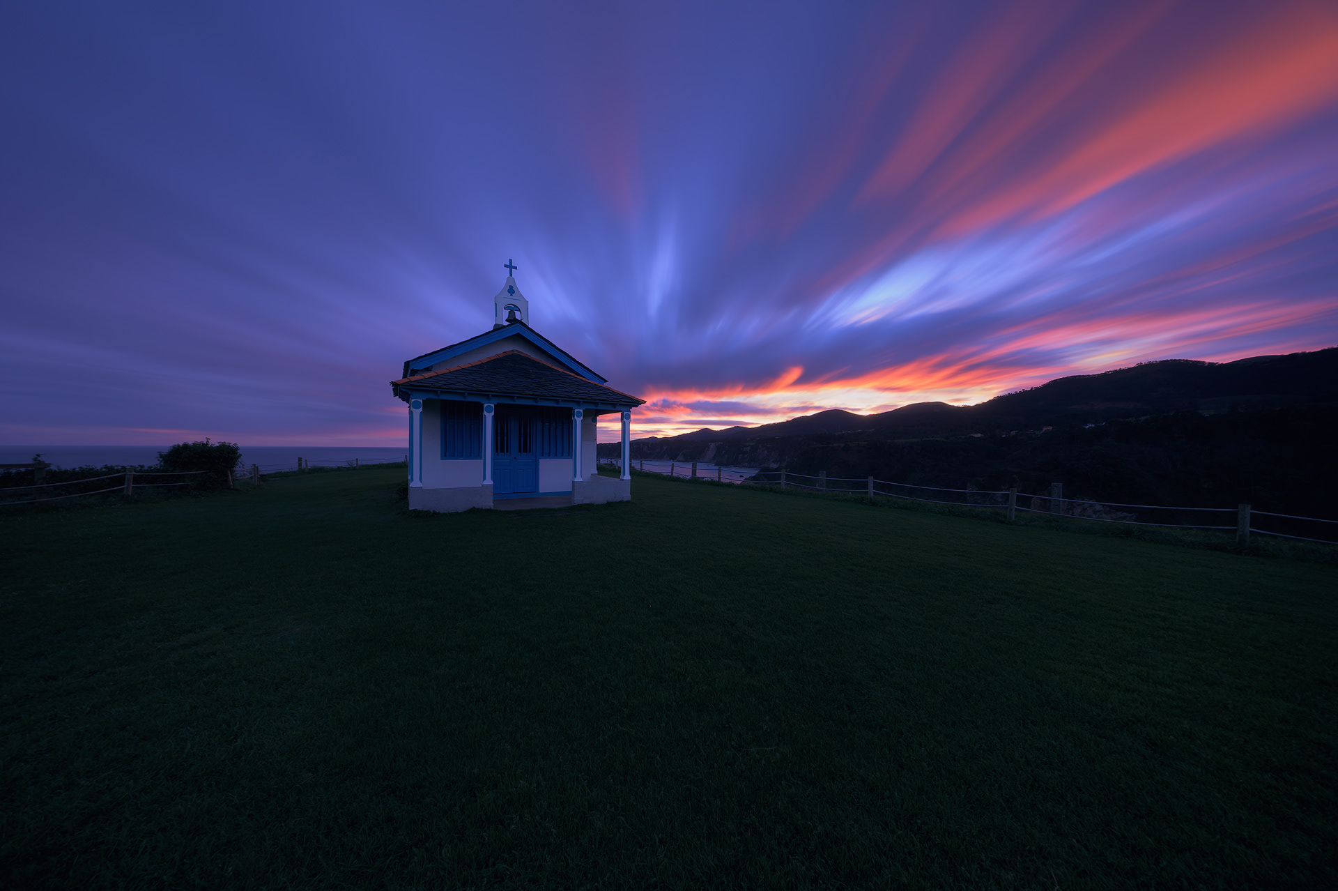Amanecer en La Regalina. Una pequeña ermita blanca con detalles en azul se encuentra en el centro de un campo verde. El cielo está lleno de nubes coloridas, con tonos de azul, púrpura y naranja, debido al amanecer. El viento mueve las nubes, creando un efecto visual impresionante. La ermita destaca claramente contra el fondo del cielo y el paisaje, disfrutando de los primeros rayos de sol del día