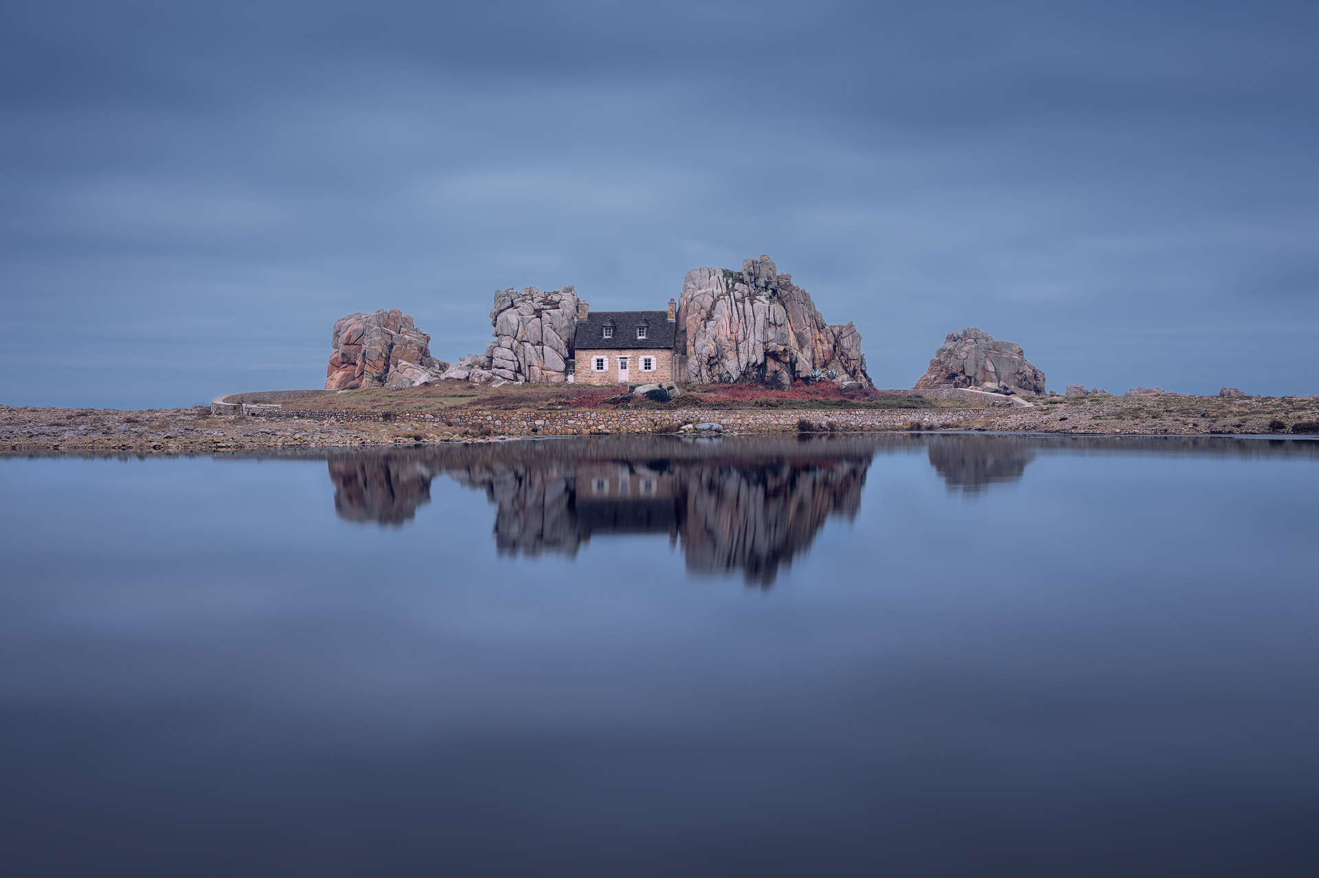 Fotografía de Castel Meur, la icónica casa de piedra encajada entre dos grandes rocas de granito en el Gouffre de Plougrescant, en la costa norte de Bretaña, Francia. En primer plano, el agua refleja suavemente la casa y las rocas, logrando un efecto de calma gracias a una técnica de larga exposición. El cielo nublado crea un ambiente dramático, mientras los tonos naturales del paisaje destacan con armonía.