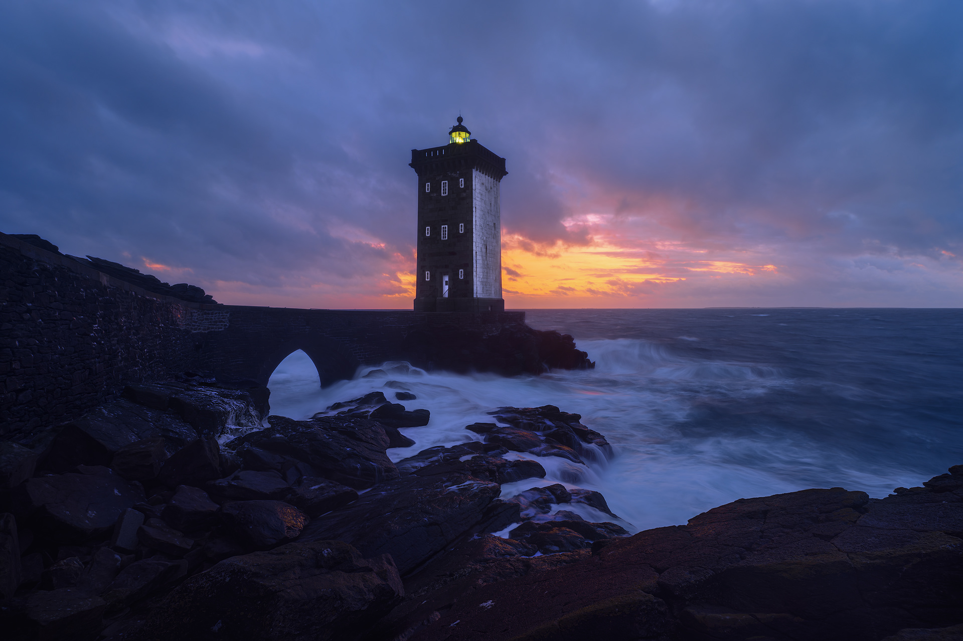 Fotografía del Phare de Kermorvan al atardecer. El faro de piedra, de forma cuadrada, se alza sobre un paisaje rocoso mientras las olas rompen contra las rocas. El cielo muestra tonos de azul oscuro con destellos anaranjados y rosados en el horizonte. La luz del faro brilla suavemente, destacando en la escena costera.