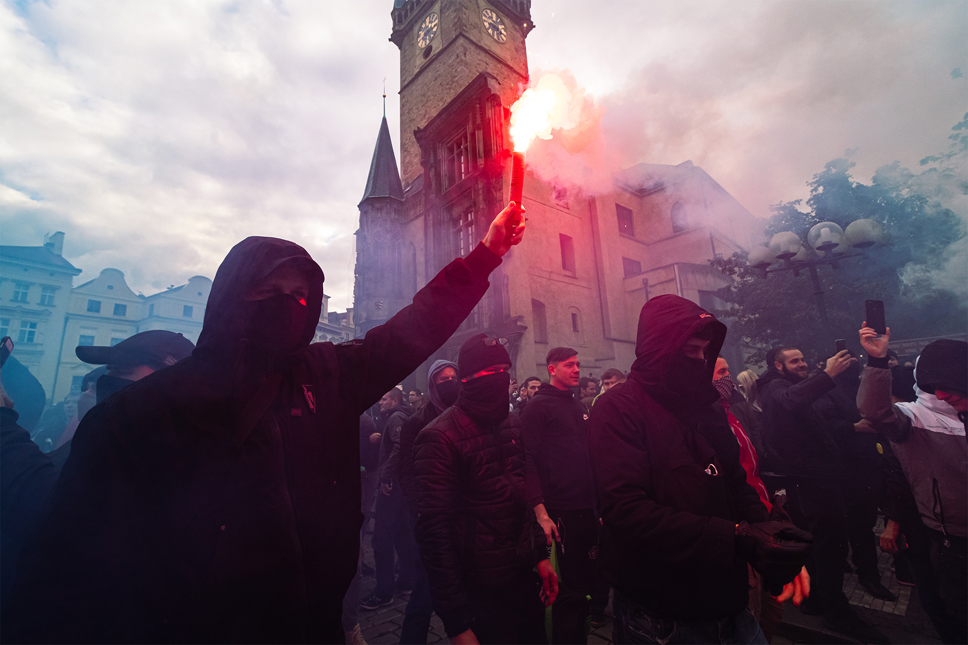 Reportážní fotografie zahaleného demonstranta, který drží červenou dýmovinice při protivládní demonstraci na Staroměstském náměstí v Praze