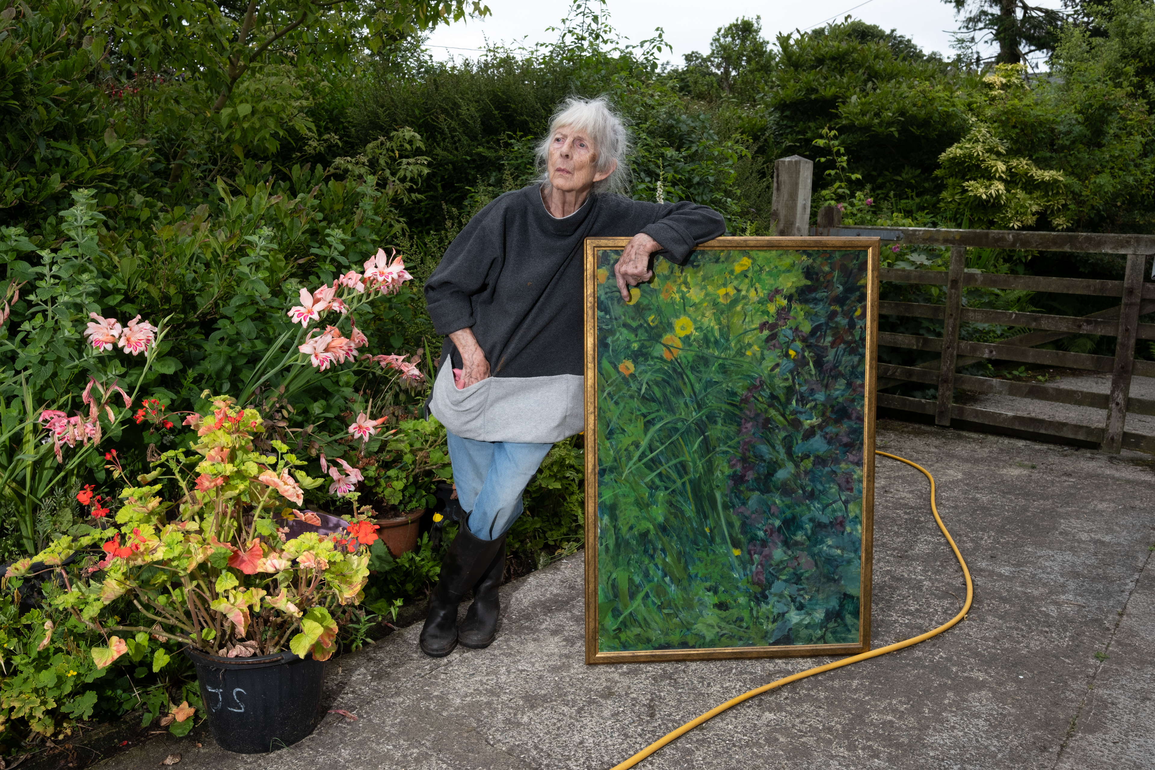 The is an image of Veronica Knowles (June 2025) - Veronica Knowles is an accomplished fine artist, gardener, and bird watcher. She tends to her garden in Sir Fon (Anglesey) - paints her garden, and feeds a myriad of birds that visit her garden. She also bakes amazing shortbread. Here she poses with one of her garden paintings whilst watching Wood Pigeons feeding in the distance.