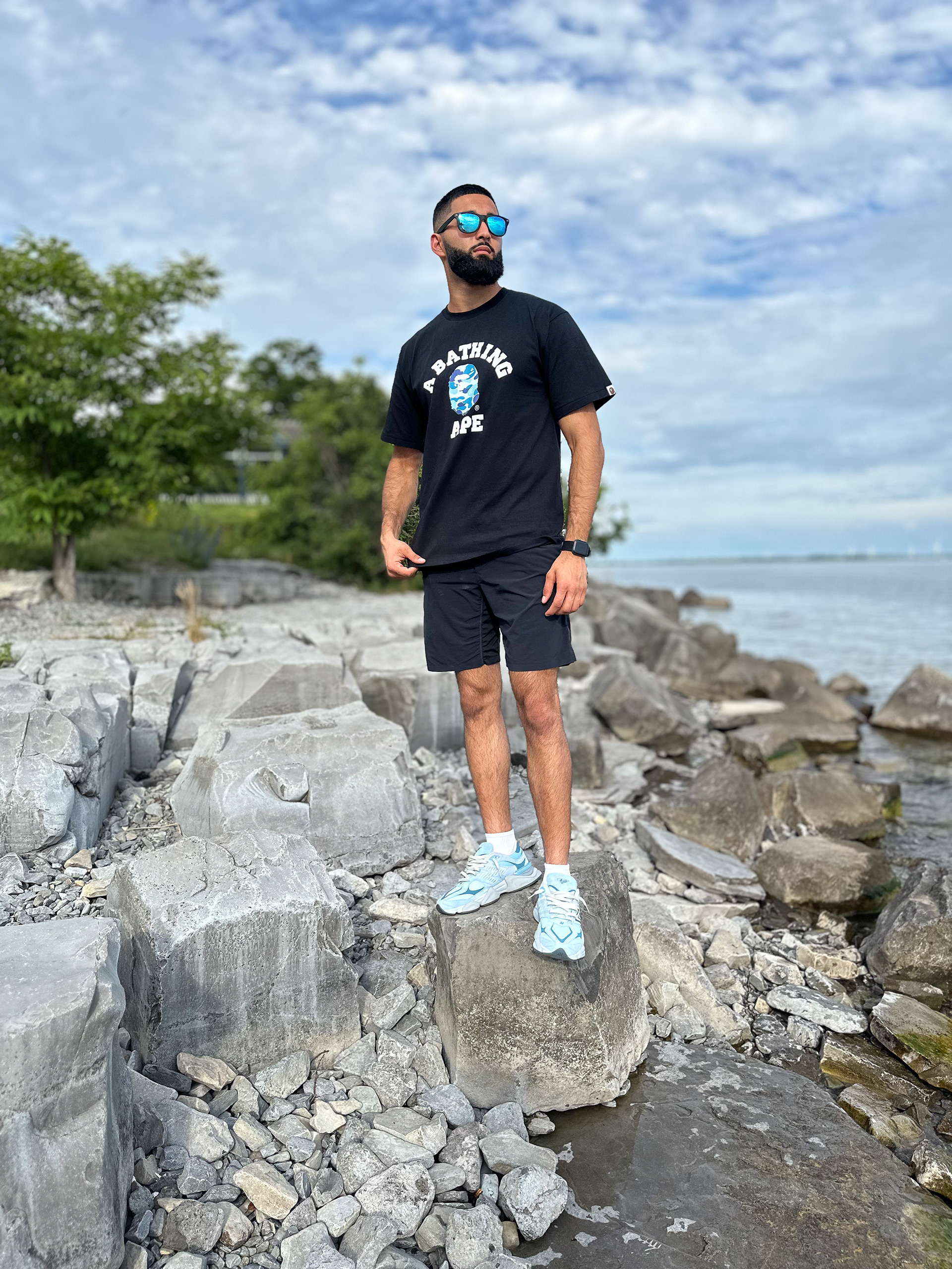 Muhammad Abbasi | Lifestyle | Black athletic graphic t-shirt, black shorts, light blue sneakers, standing on rocky shoreline