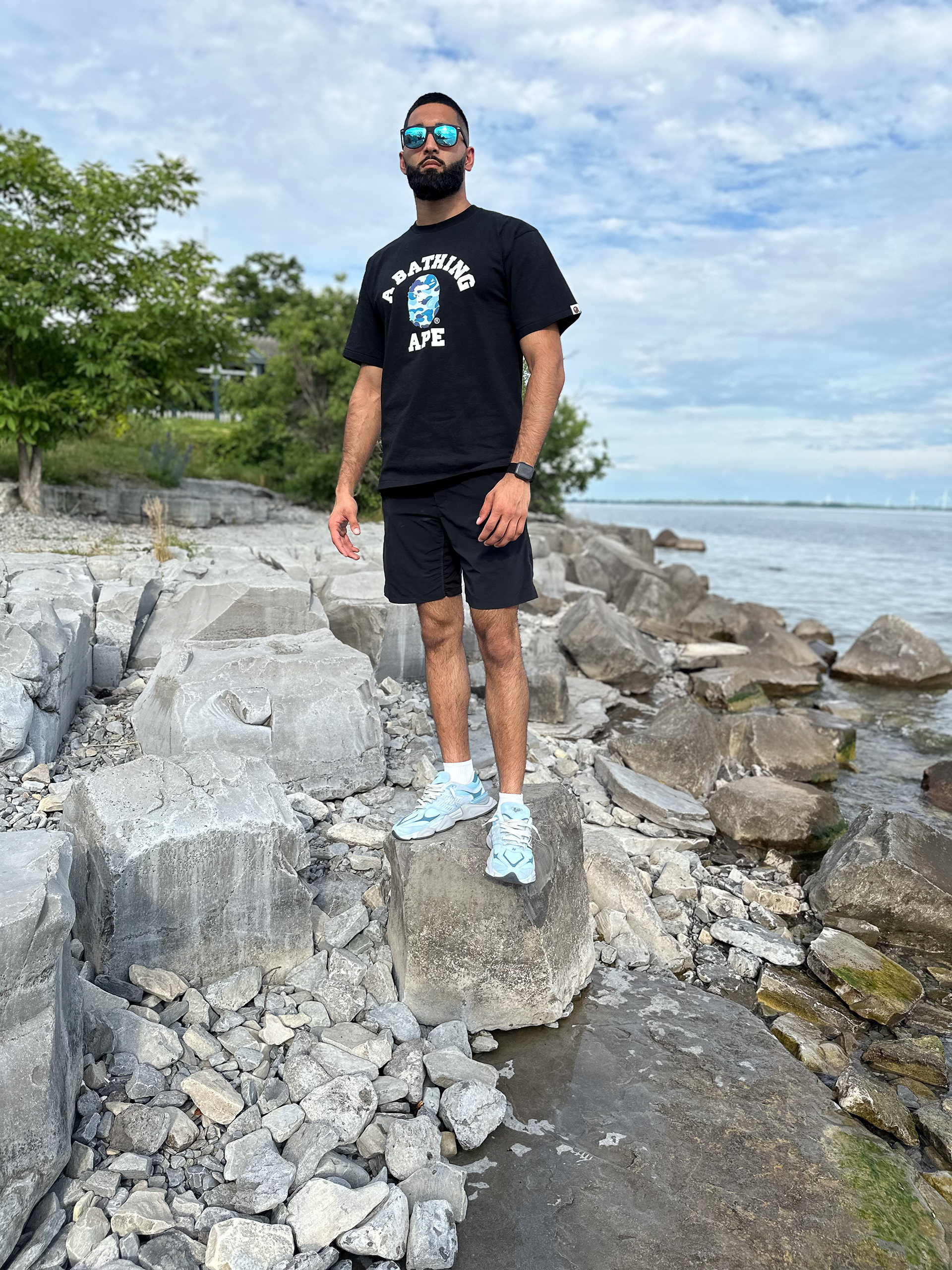 Muhammad Abbasi | Lifestyle | Black graphic t-shirt, black shorts, light blue sneakers, facing camera on lakefront rocks