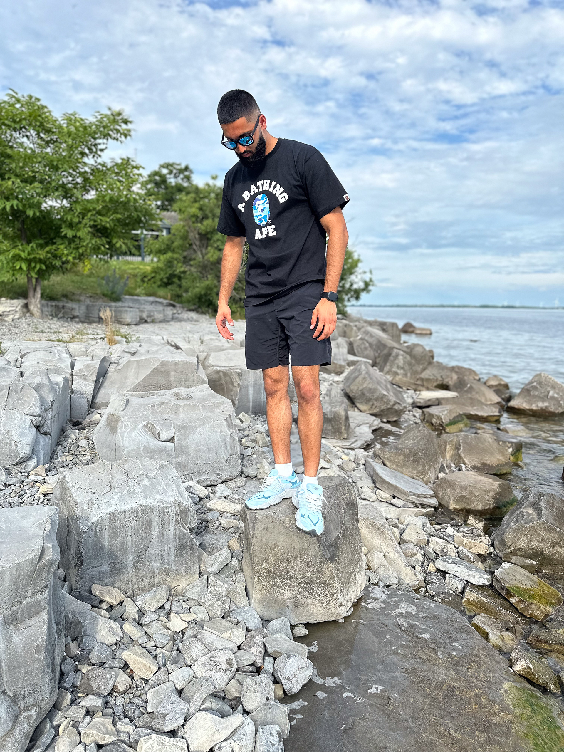 Muhammad Abbasi | Lifestyle | Black graphic t-shirt, black shorts, light blue sneakers, looking down while standing on rock