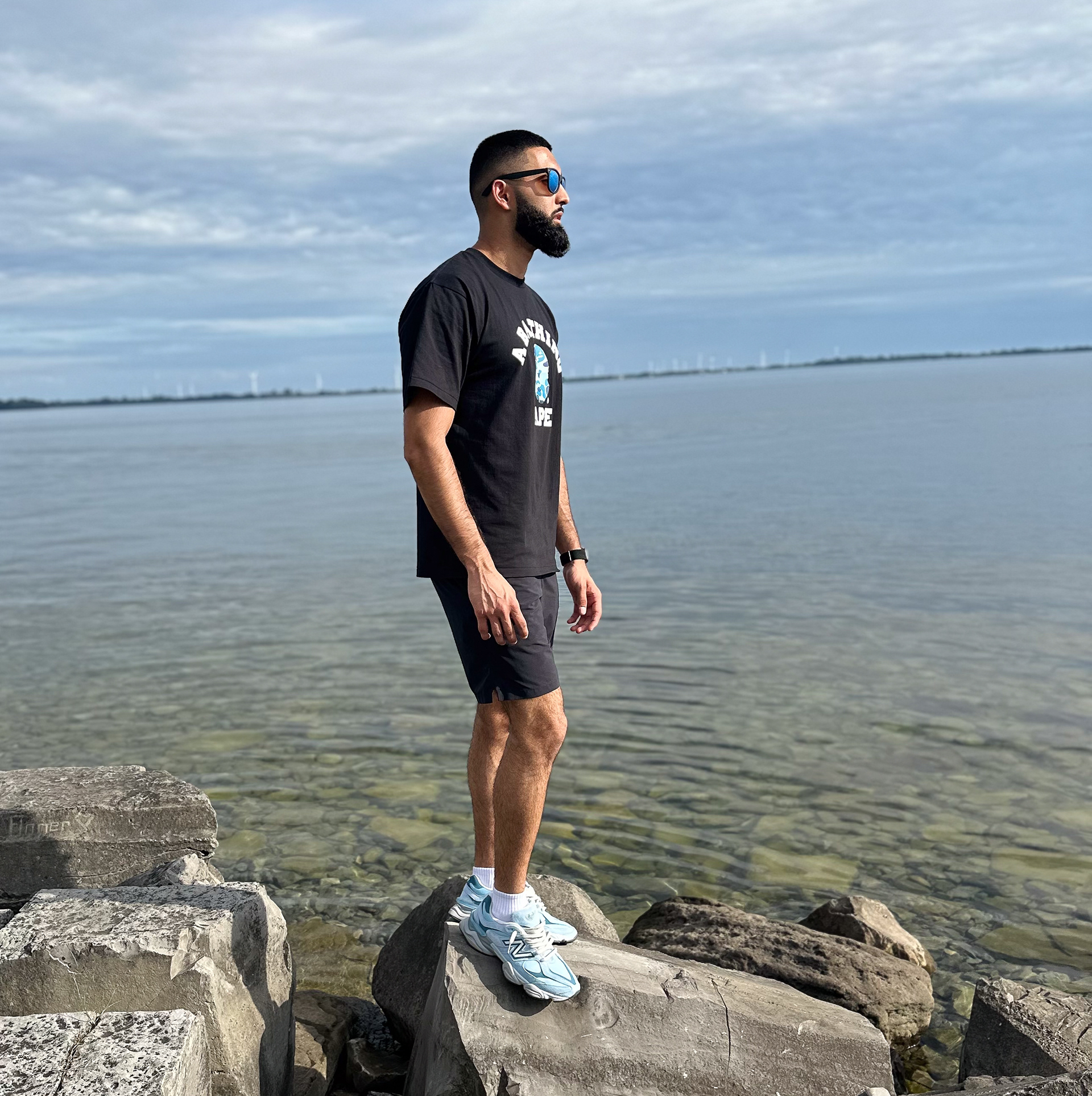 Muhammad Abbasi | Lifestyle | Black graphic t-shirt, black shorts, light blue sneakers, standing on rocks with water background