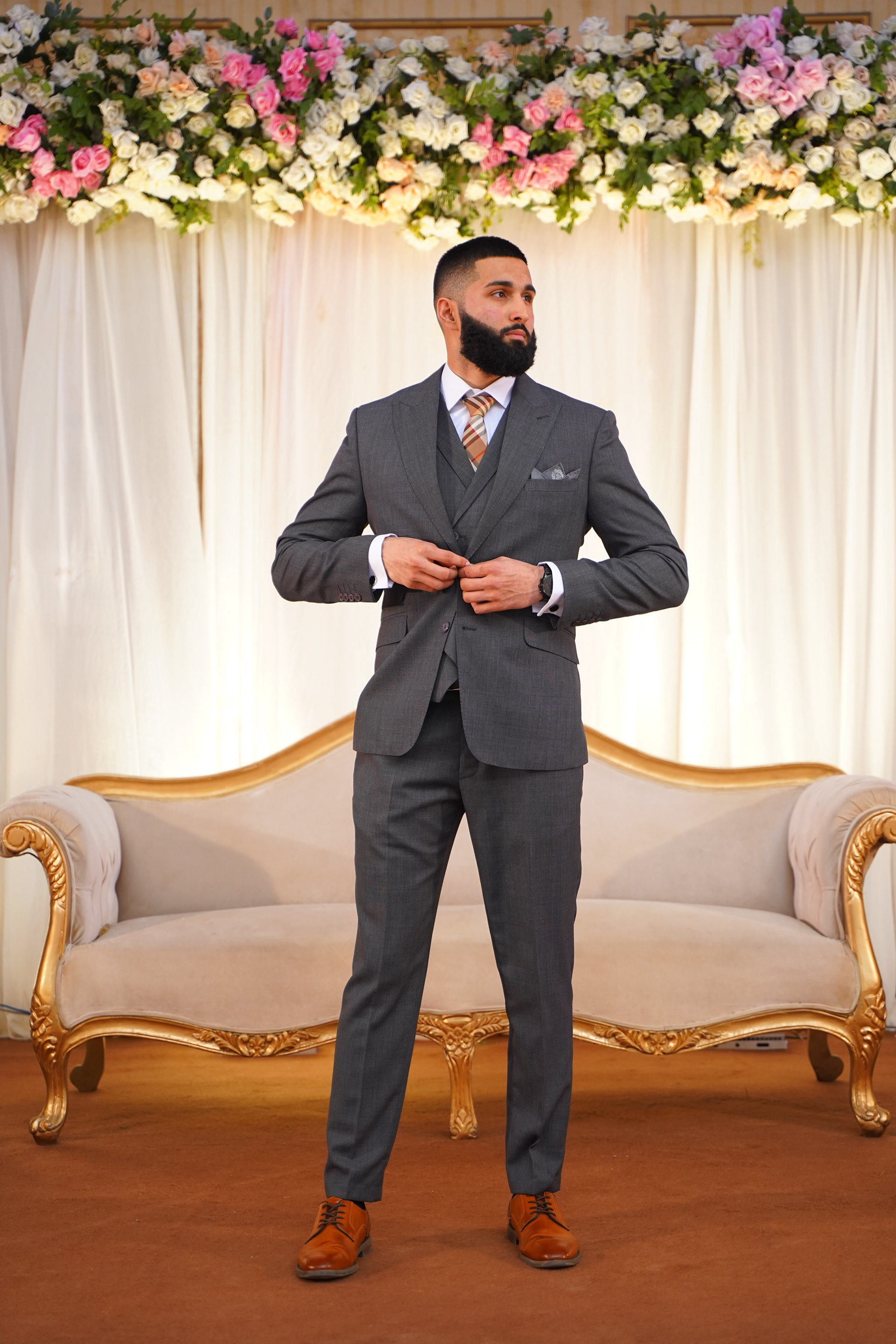 Muhammad Abbasi | Formal Wear | Charcoal three-piece suit with striped tie, adjusting jacket in front of floral backdrop