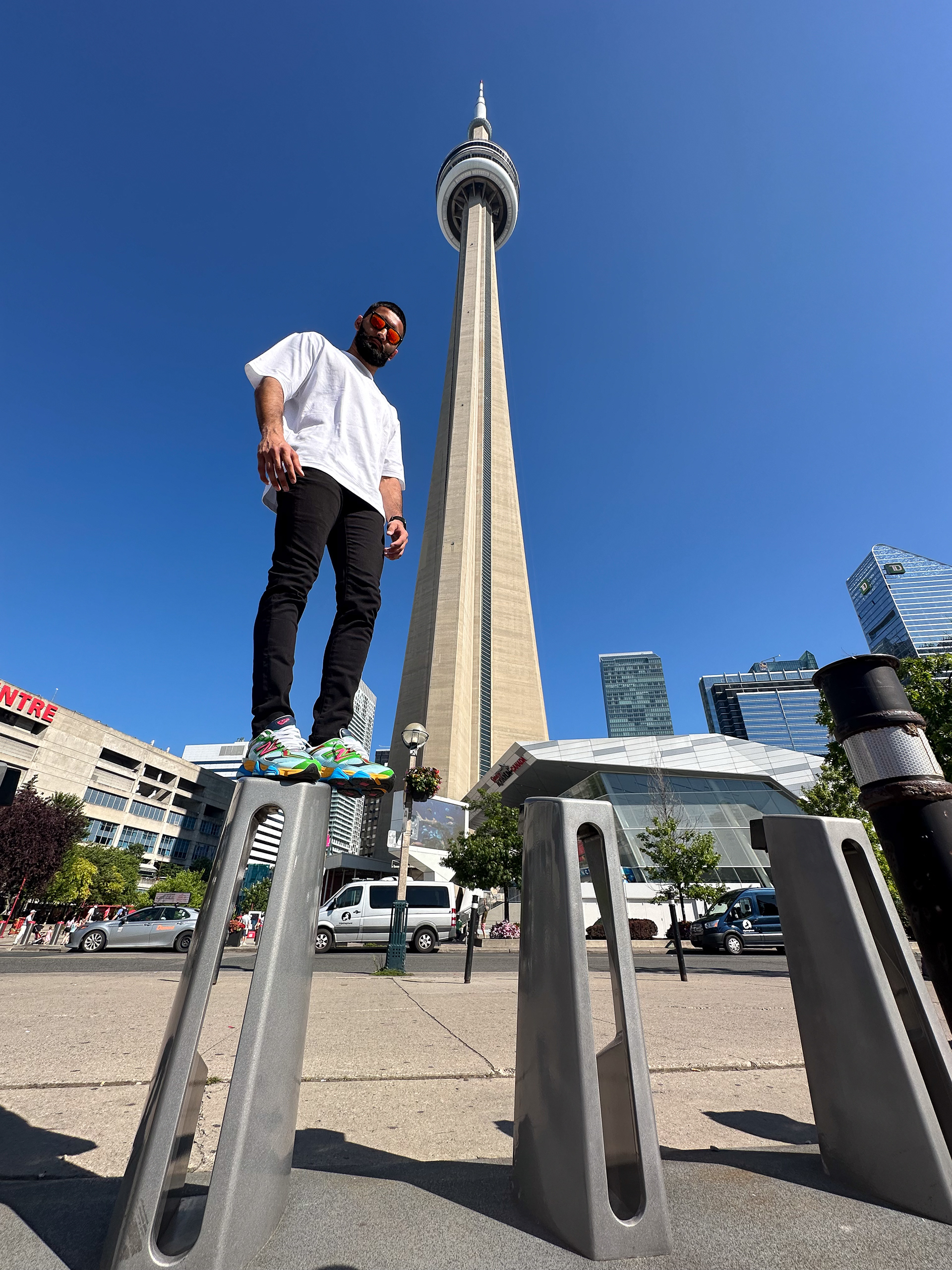 Muhammad Abbasi | Lifestyle | White oversized t-shirt, black jeans, colorful sneakers, standing on bike rack with CN Tower in background