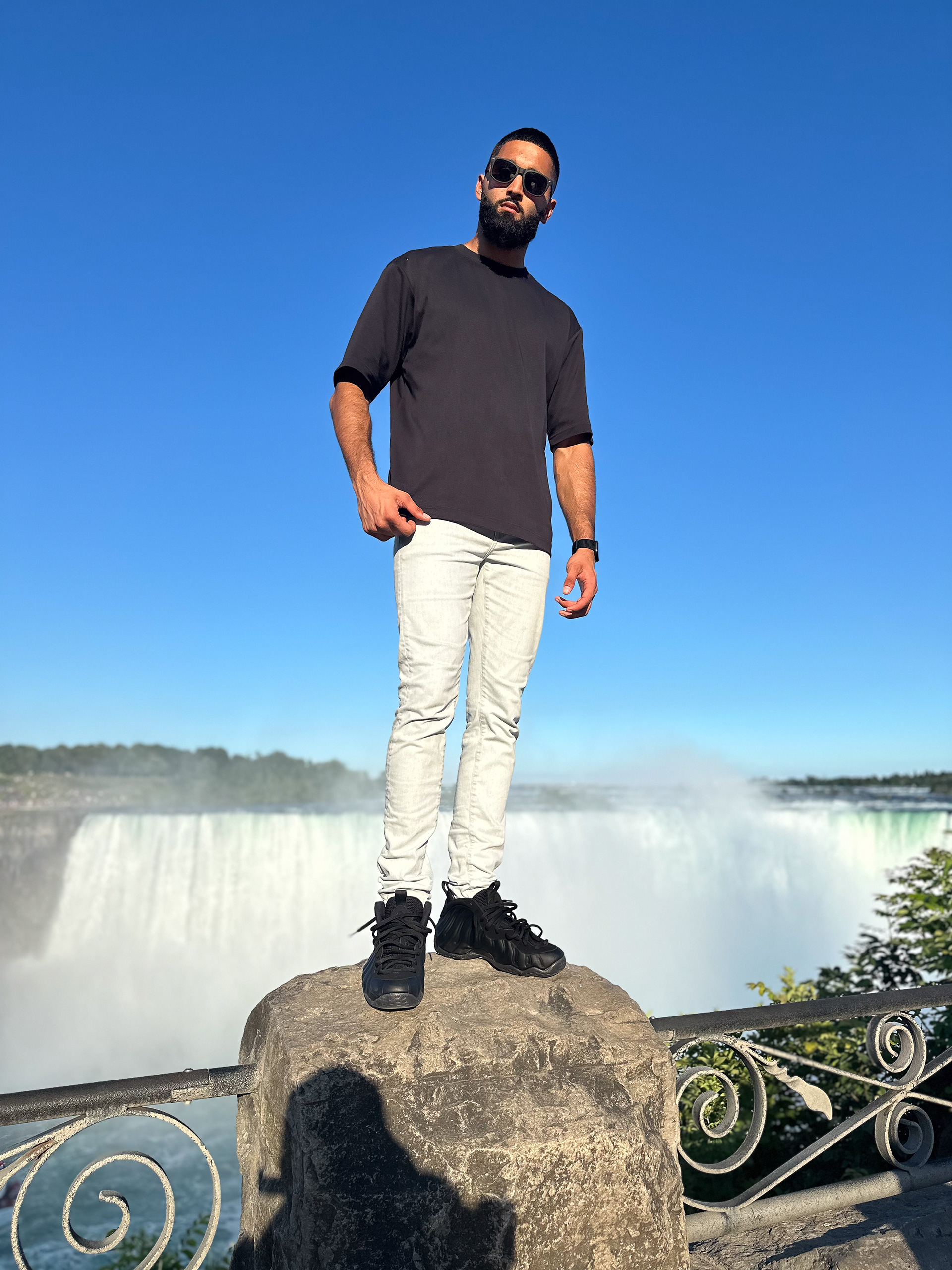 Muhammad Abbasi | Lifestyle | Black t-shirt, light blue jeans, black sneakers, standing on rock with Niagara Falls in background