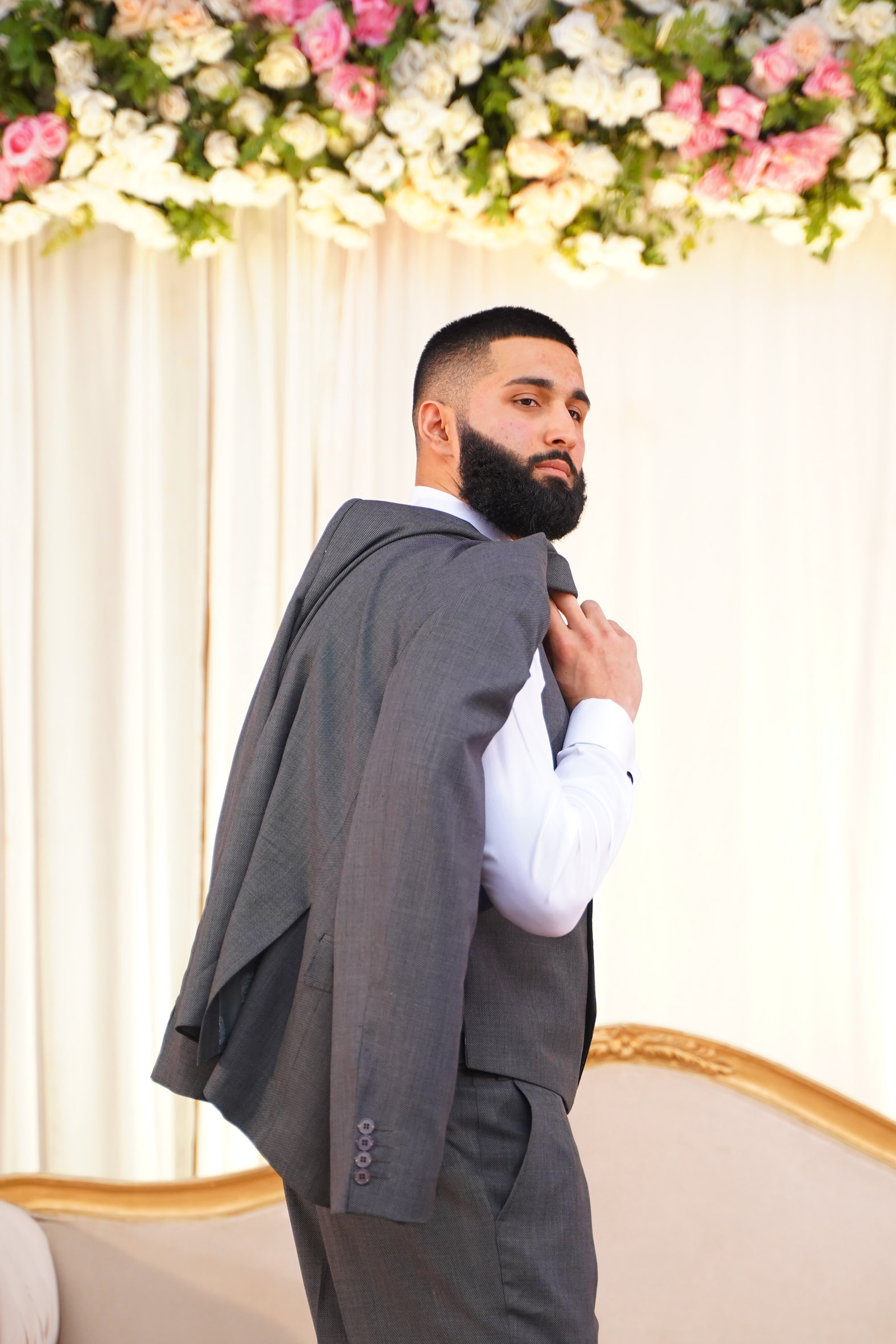 Muhammad Abbasi | Formal Wear | Charcoal three-piece suit with striped tie, holding jacket over shoulder facing away from camera