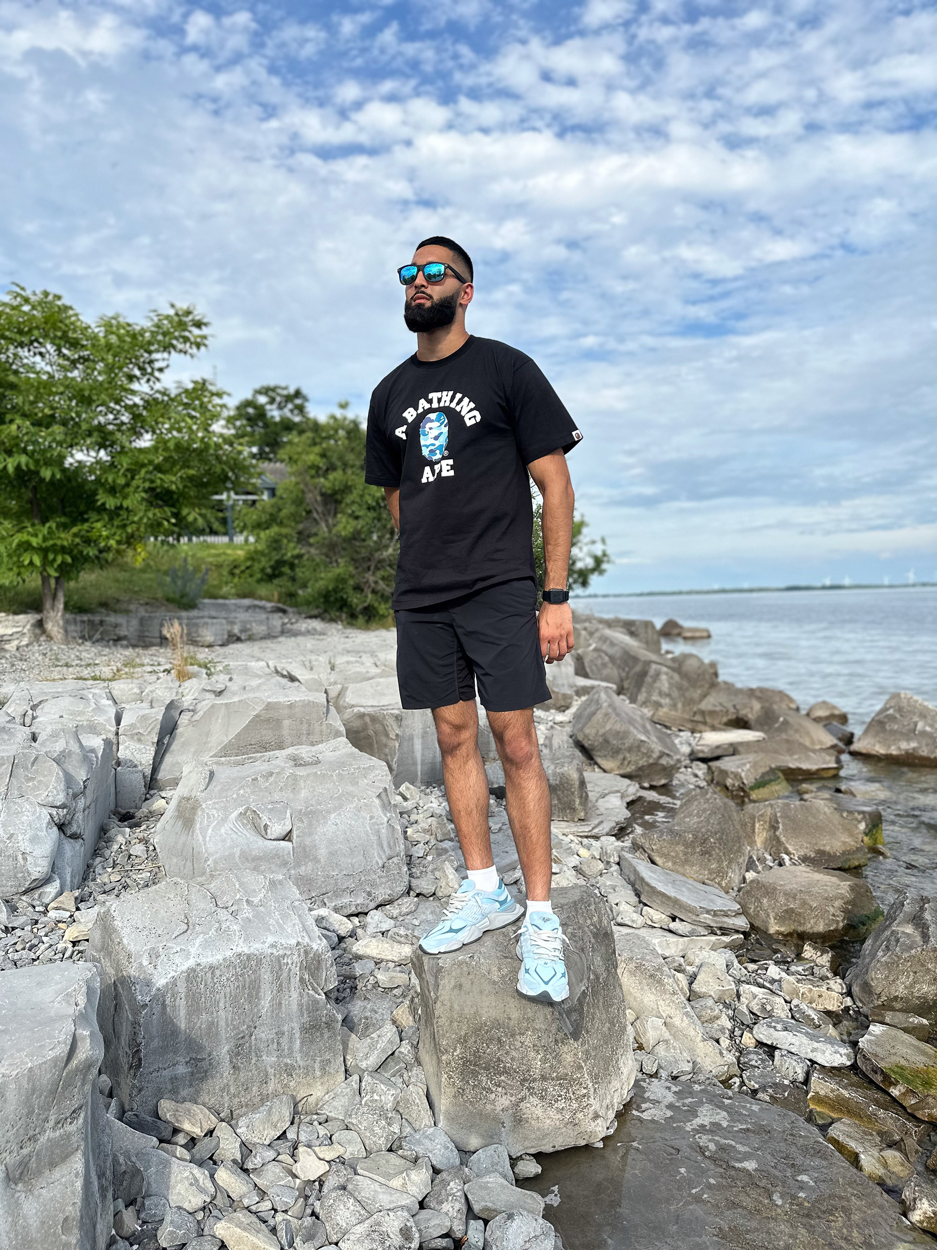 Muhammad Abbasi | Lifestyle | Black graphic t-shirt, black shorts, light blue sneakers, standing on rocks by lake