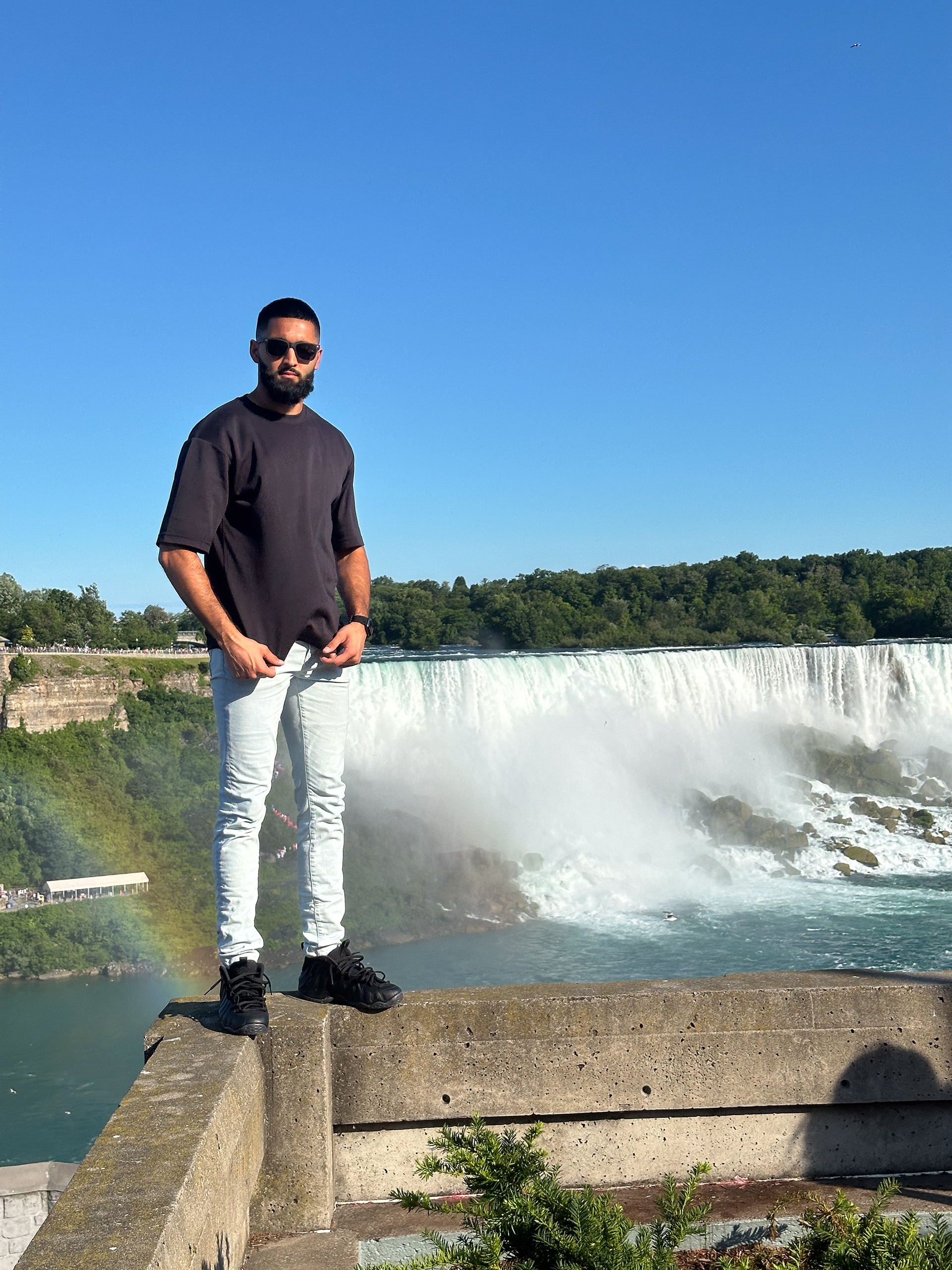 Muhammad Abbasi | Lifestyle | Black t-shirt, light blue jeans, black sneakers, standing on ledge with waterfall behind