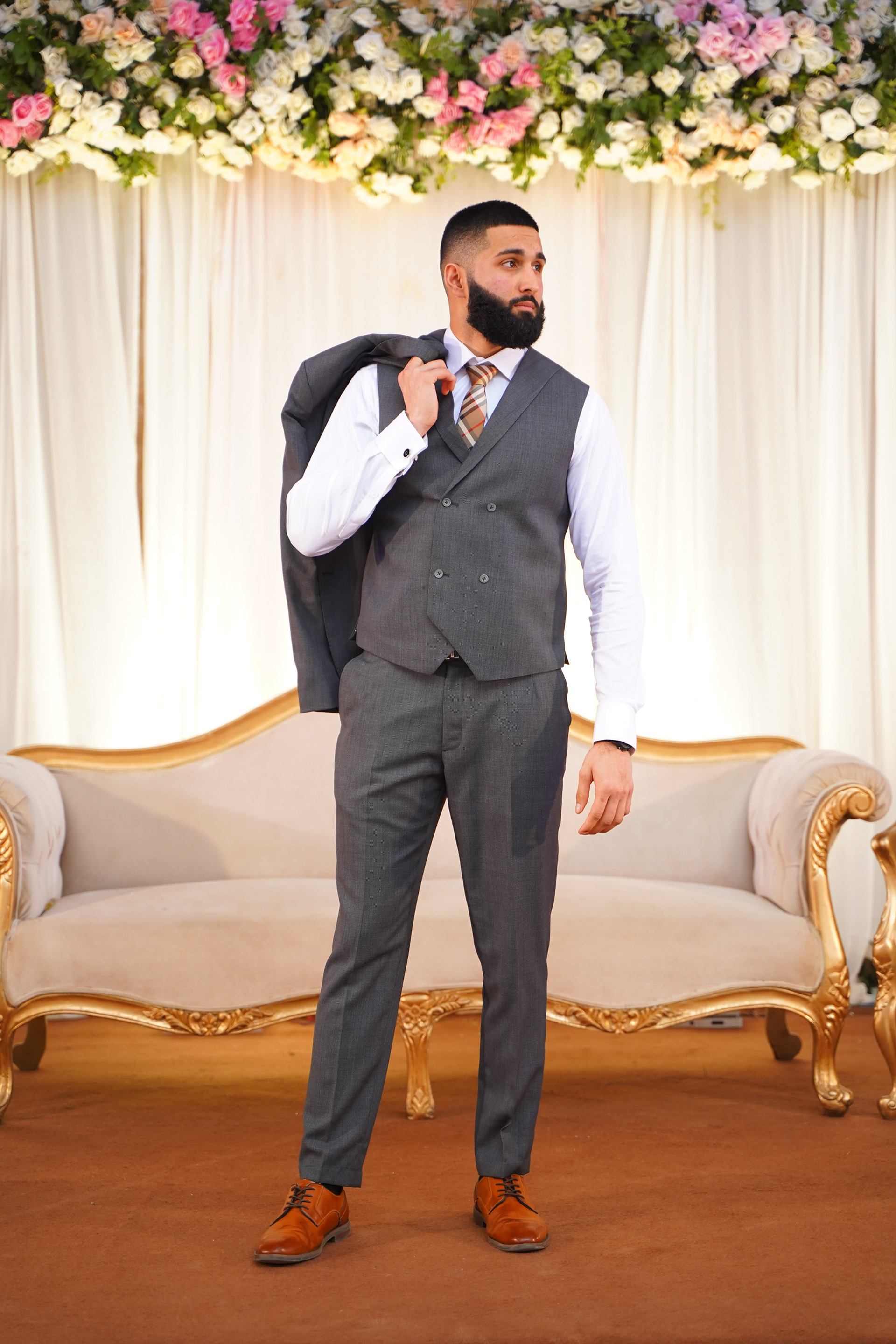 Muhammad Abbasi | Formal Wear | Charcoal three-piece suit with striped tie, jacket over shoulder in front of floral backdrop