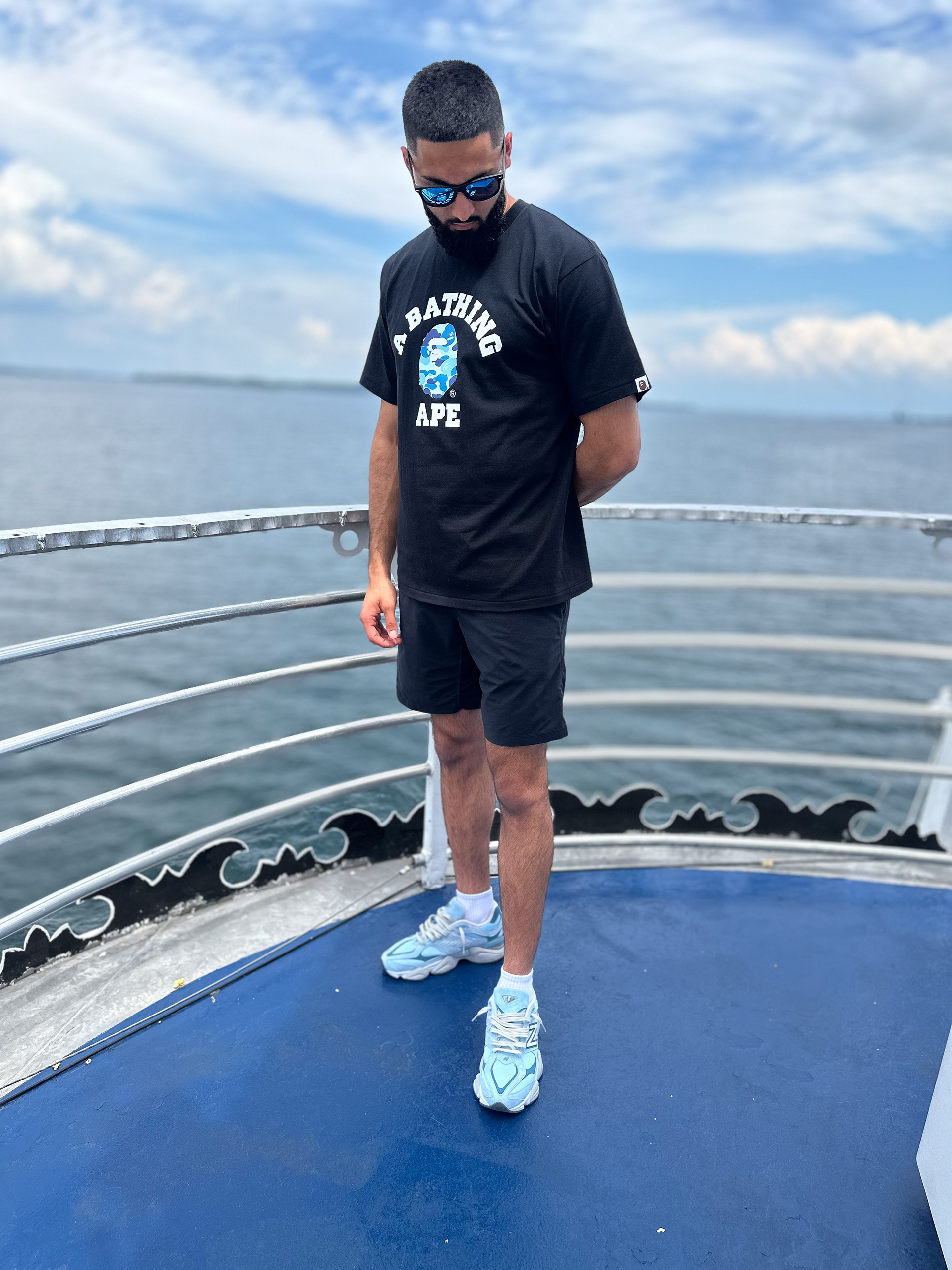 Muhammad Abbasi | Lifestyle | Black graphic t-shirt, black shorts, light blue sneakers, looking down on boat deck by water