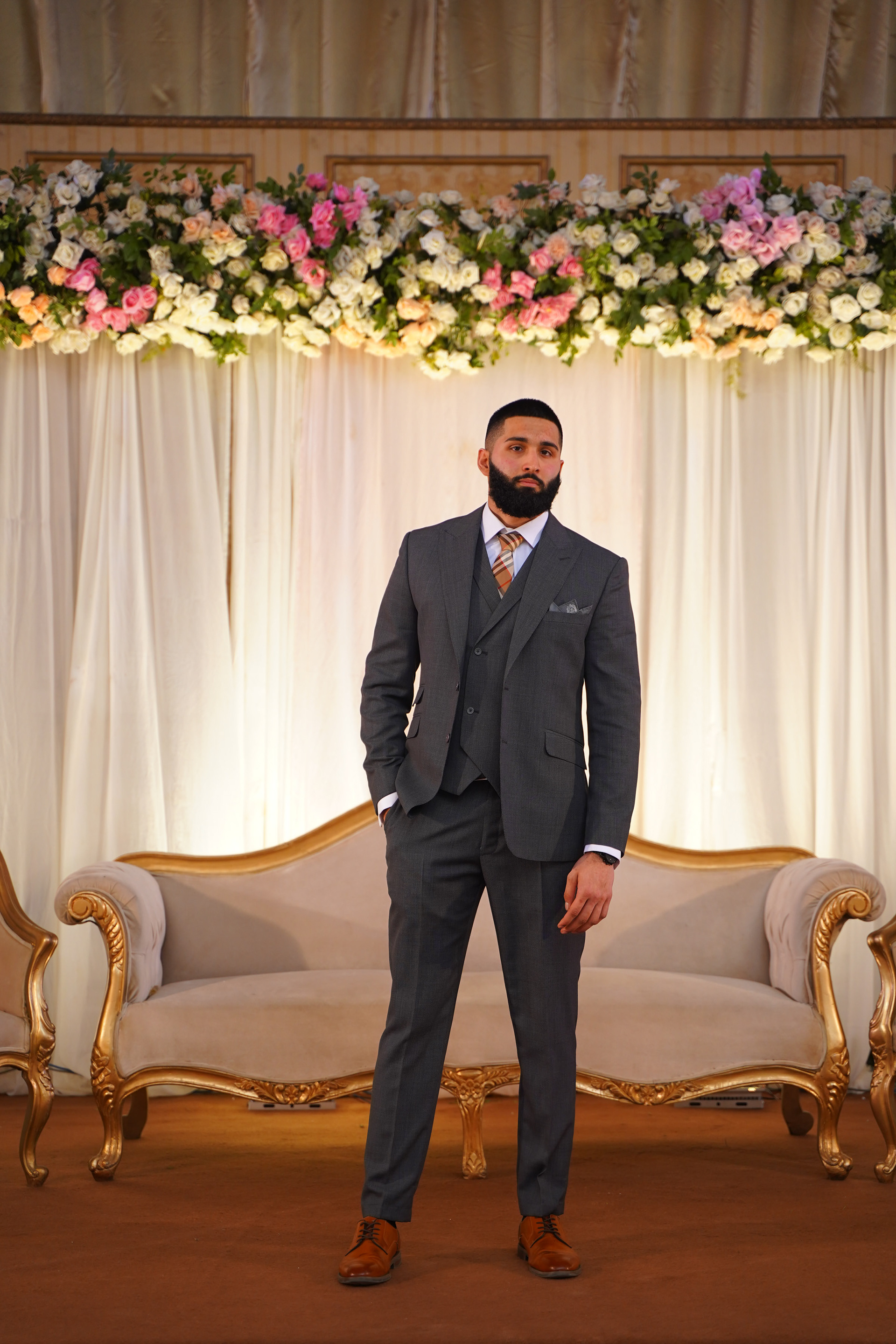 Muhammad Abbasi | Formal Wear | Charcoal three-piece suit with striped tie, standing in front of floral backdrop with hand in pocket