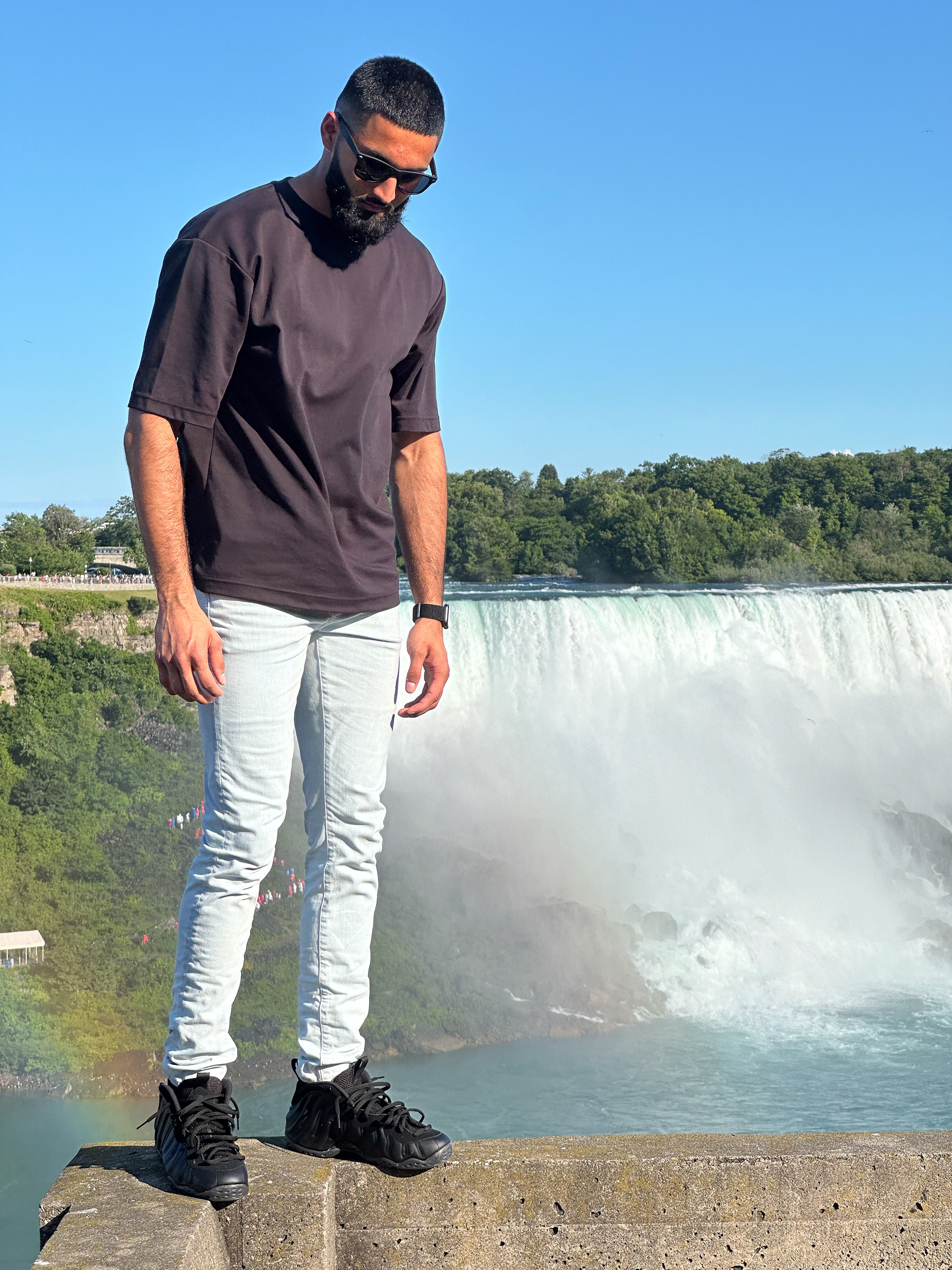 Muhammad Abbasi | Lifestyle | Black t-shirt, light blue jeans, black sneakers, looking down with Niagara Falls in background