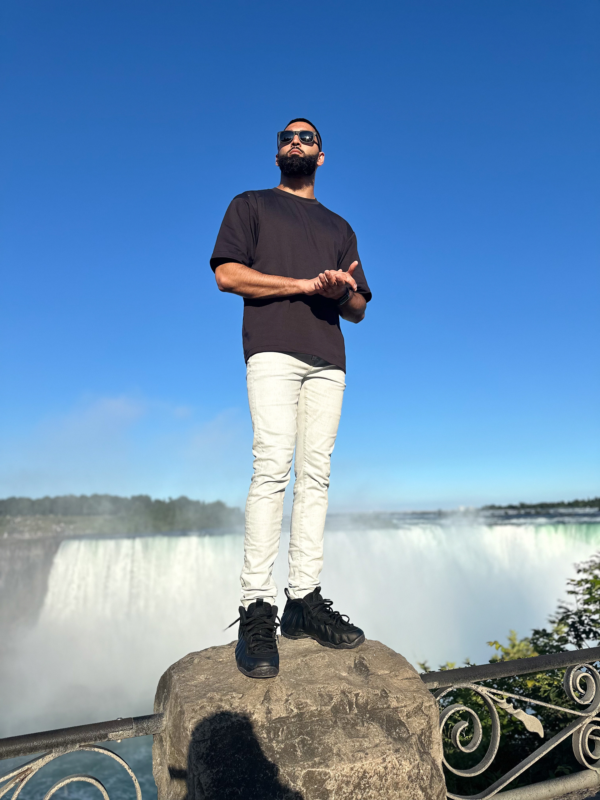Muhammad Abbasi | Lifestyle | Black t-shirt, light blue jeans, black sneakers, standing on rock with Niagara Falls in background