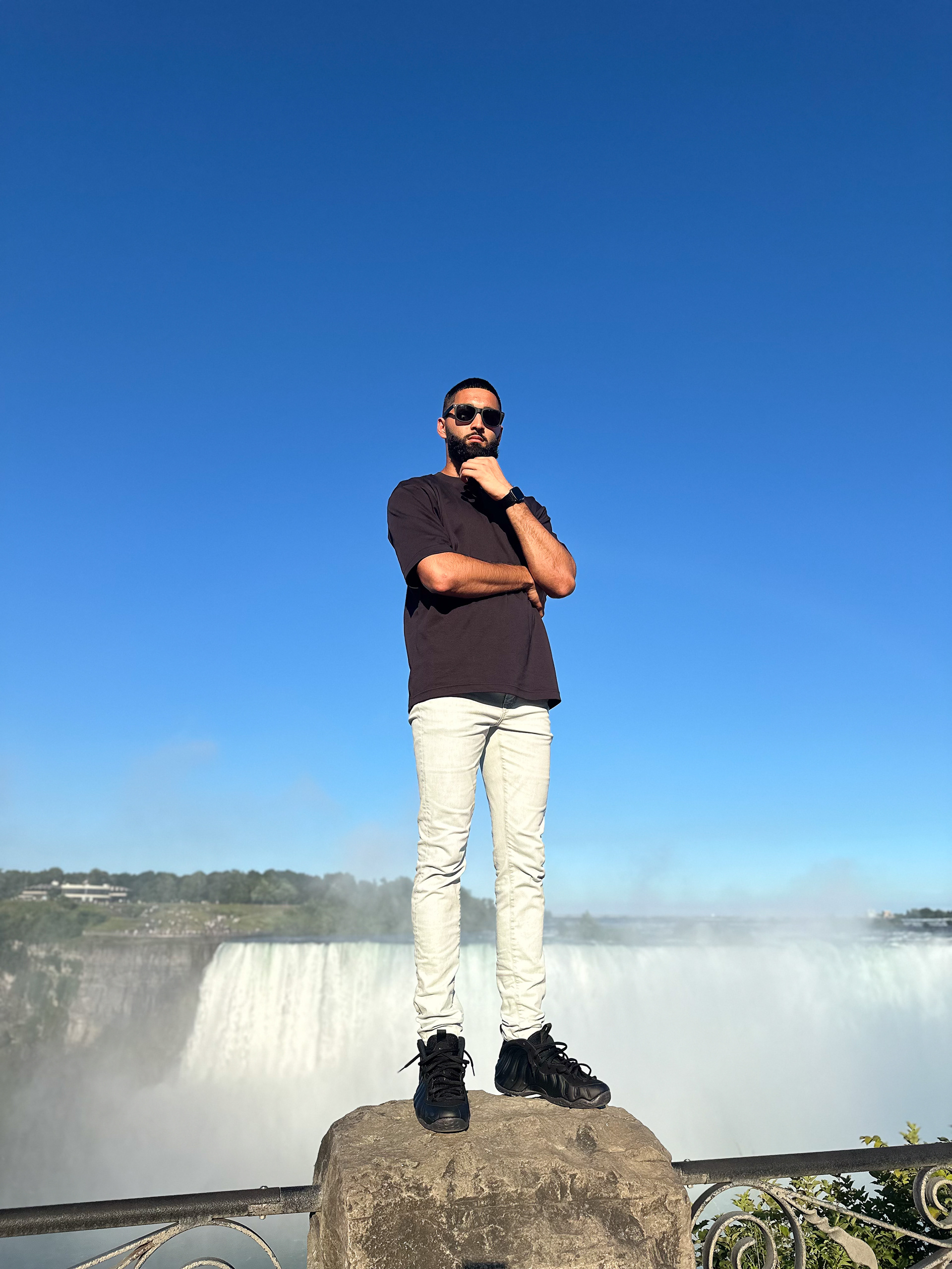 Muhammad Abbasi | Lifestyle | Black t-shirt, light blue jeans, black sneakers, arms crossed, standing on rock with Niagara Falls in background