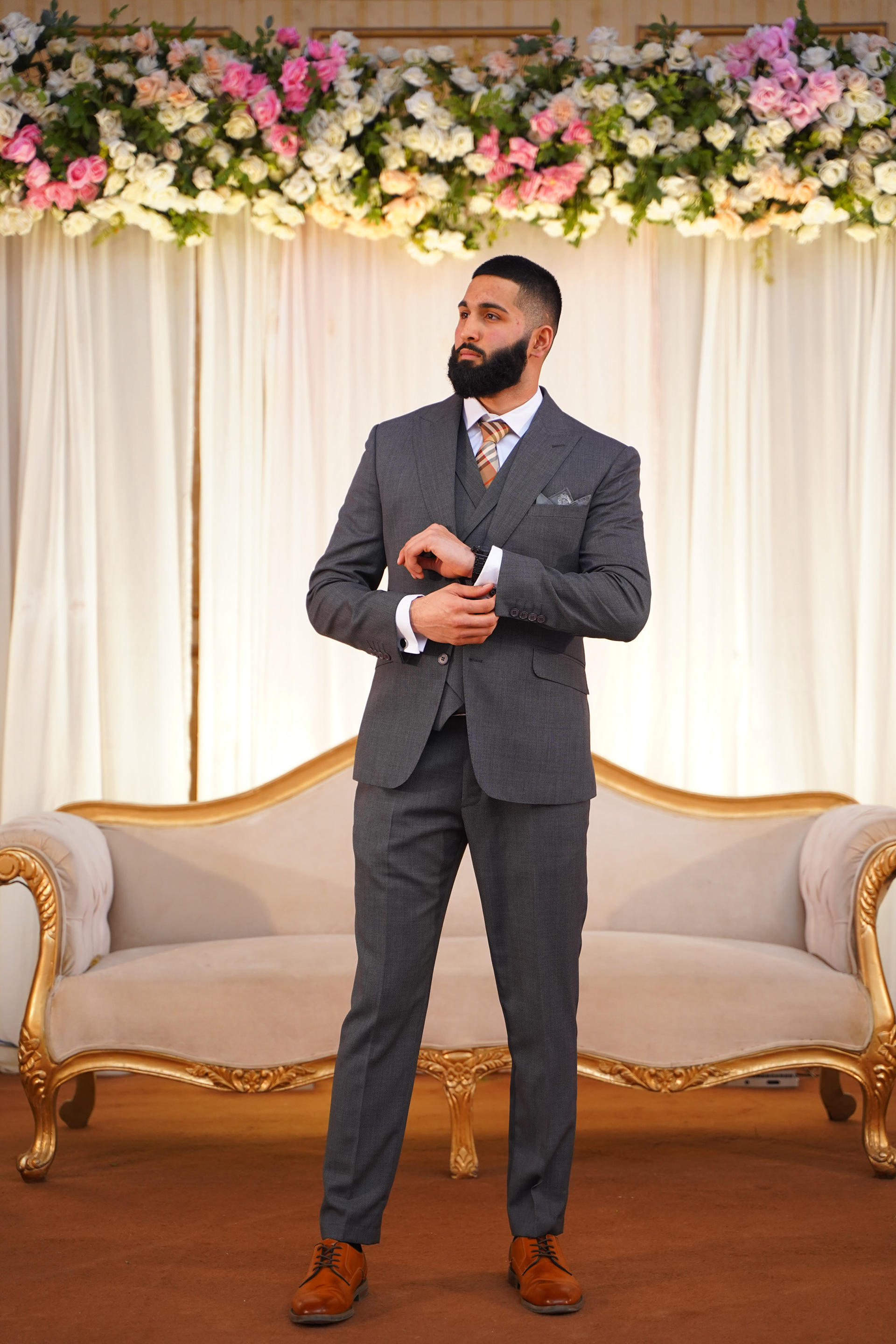 Muhammad Abbasi | Formal Wear | Charcoal three-piece suit with striped tie, buttoning cuff link in front of floral backdrop