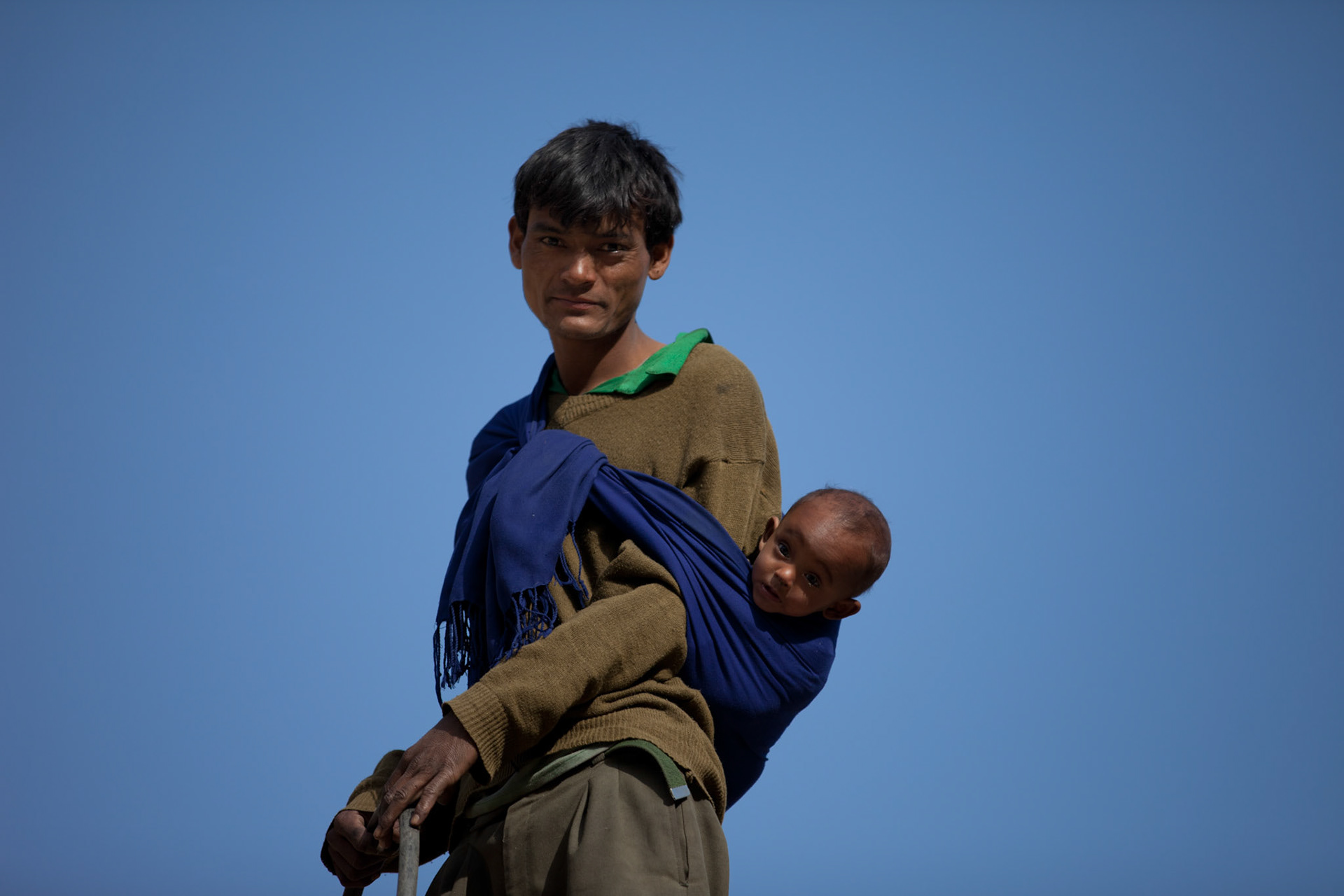 A man with a baby on his back stands with a blue sky behind. The Karnali area of Nepal in the mid western hills and along the Chinese border is one of the most food insecure regions in Asia. The World Food Program estimate 75% of the children in the district have stunted growth and up to 50% are malnourished. With an expanding population and drying climate the challenge ahead is significant. 24/04/2011, Karnali, Nepal.
