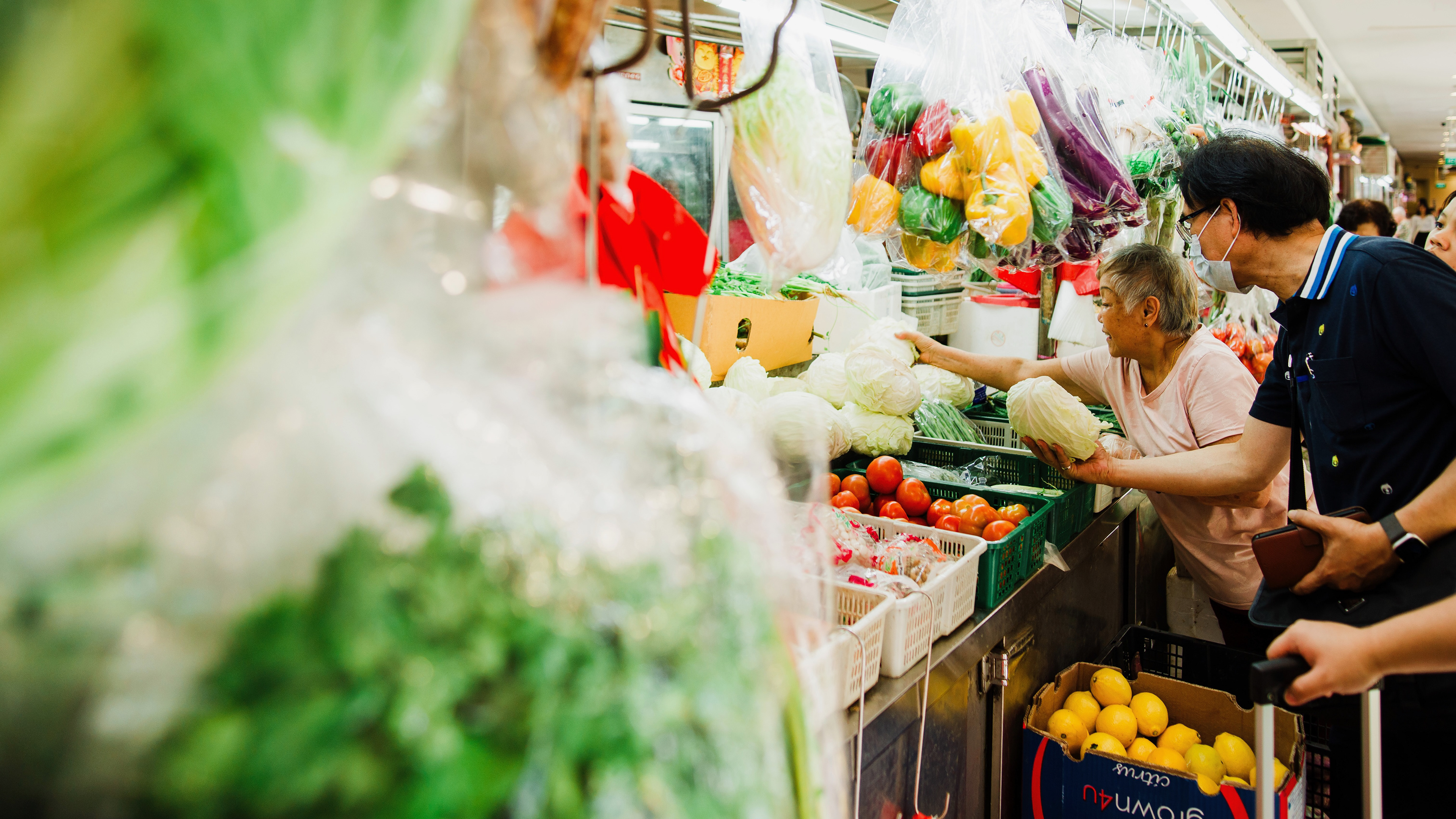 A cinematic 16:9 directorial frame of a traditional Singaporean wet market. Low-angle shot through out-of-focus foreground elements, capturing a human interaction between vendors under fluorescent lights. Industrial noir aesthetic with high-fidelity geometric lines and a voyeuristic, documentary soul.