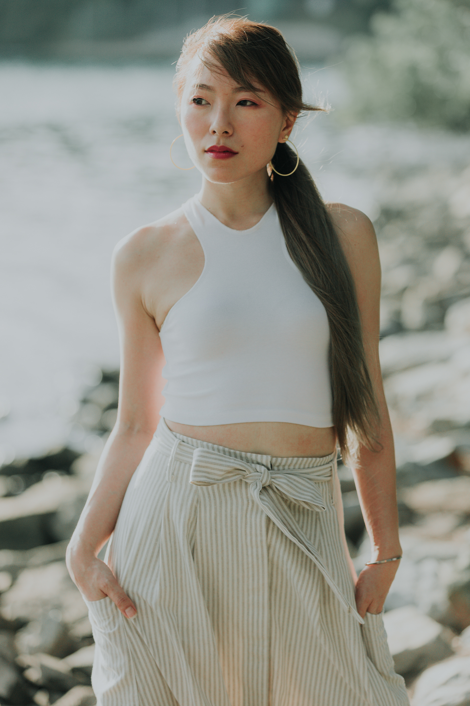 Cinematic high-fidelity portrait of a woman at a rocky shoreline; soft backlight creating an atmospheric bloom, capturing subtle human expression and environmental texture in a 16:9 directorial framing.