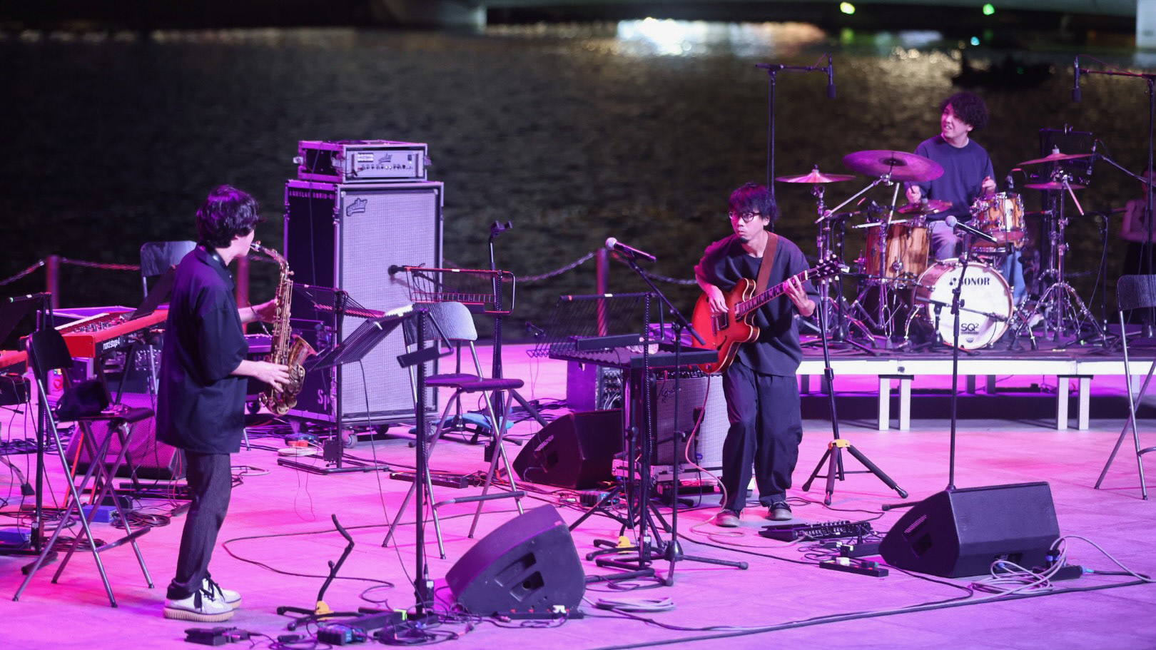A wide-angle 16:9 directorial frame of a four-piece jazz band performing on the Esplanade outdoor stage. The image documents the complete mechanical soul of the performance, including a large amplifier, drum kit, and microphone setup. Captured from an elevated audience POV, establishing the scale of the live event in Singapore.