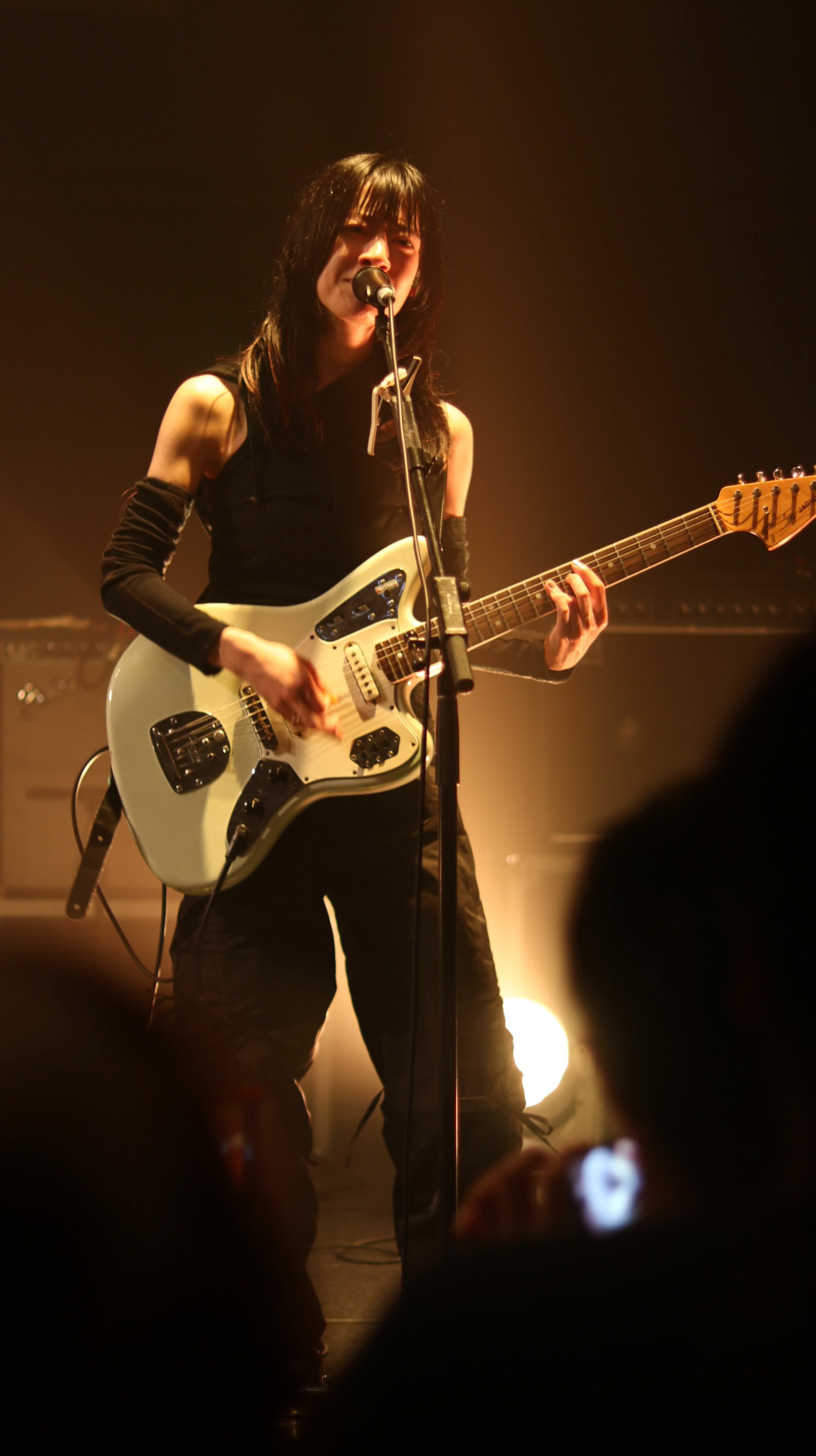 A high-fidelity directorial portrait of Hitsujibungaku guitarist Moeka Shiotsuka performing with her signature Fender Mustang. The frame captures a moment of "Active Identity" and peak delivery, with a layered foreground of audience silhouettes providing authentic energy from a "Paid Audience" POV.