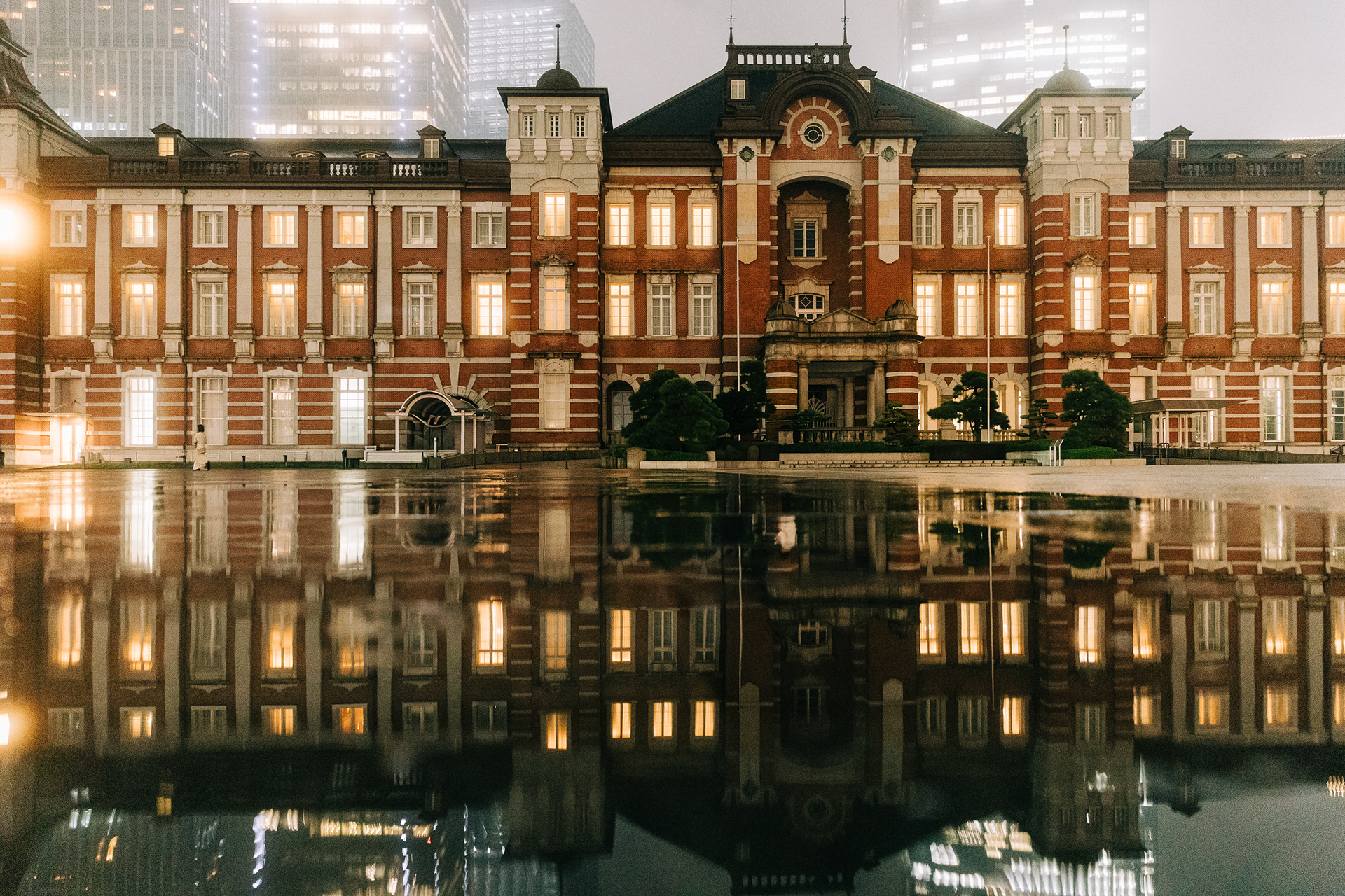 A symmetrical, high-fidelity architectural shot of Tokyo Station at night, reflected perfectly in a large rain puddle. Warm interior lighting contrasts with the cool, misty silhouettes of modern skyscrapers in the background. Stark geometric balance and cinematic atmosphere.