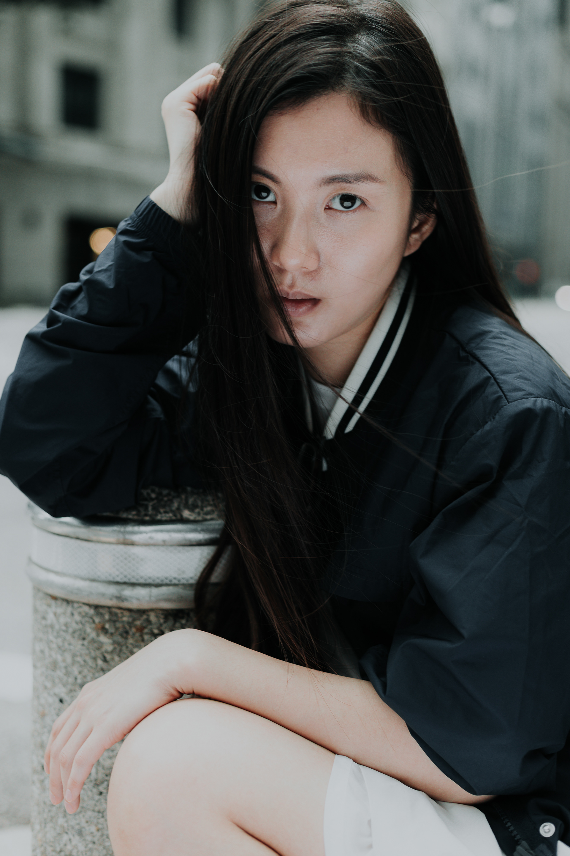 Vertical close-up portrait of a woman in a black and white bomber jacket sitting on an urban bollard, looking intensely at the camera with hair over one eye.