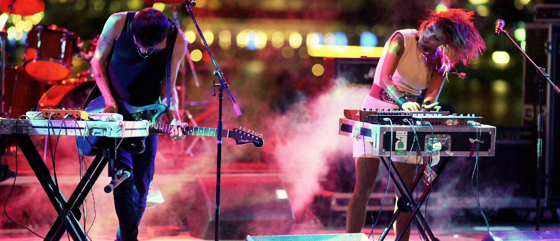 High-contrast live concert photography featuring a guitarist and singer in motion with atmospheric red and green stage lighting and haze.
