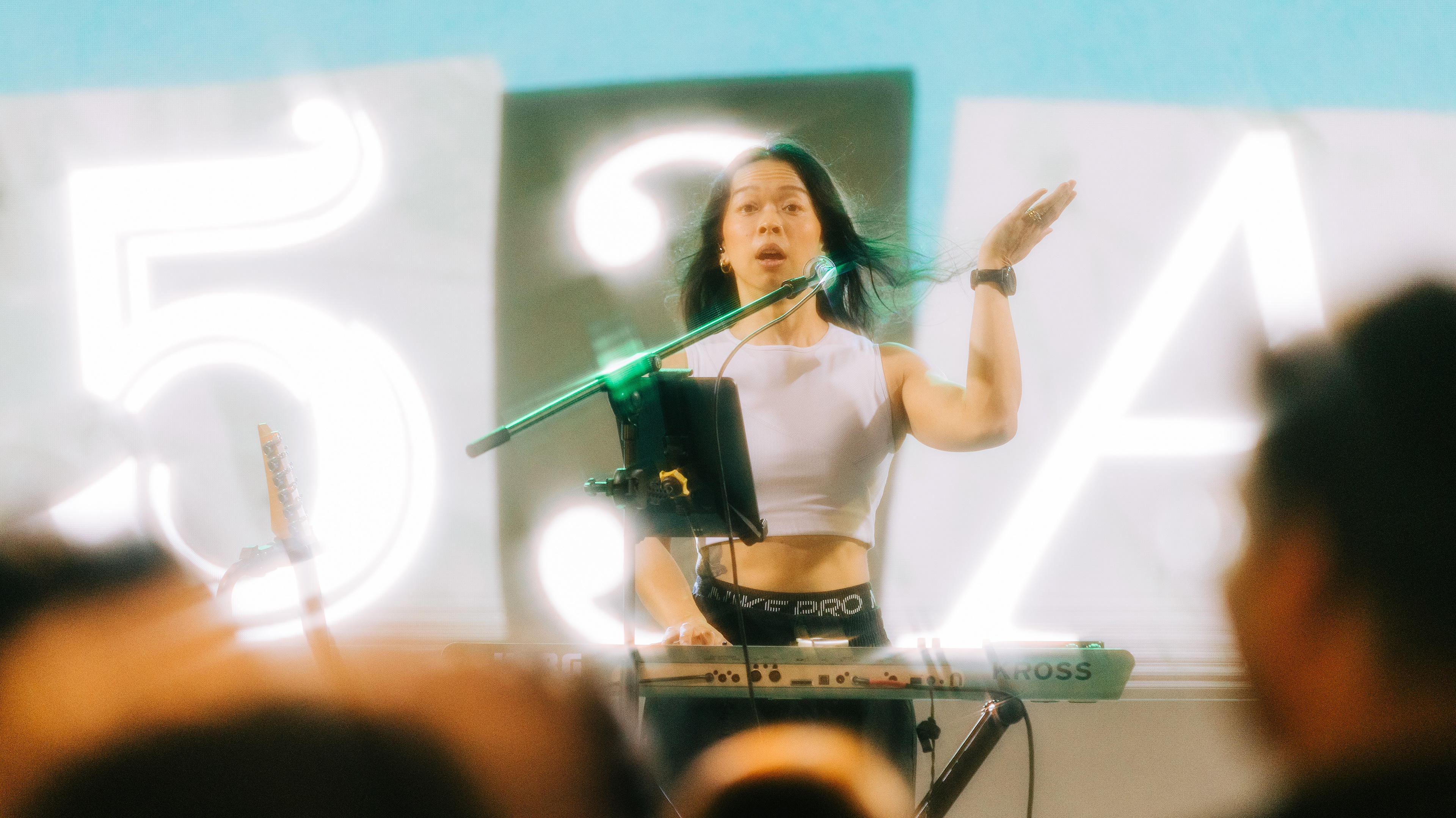 Auteur style music photography of SA53 frontwoman playing keyboard on stage, shot from behind a crowd silhouette at GastroBeats.