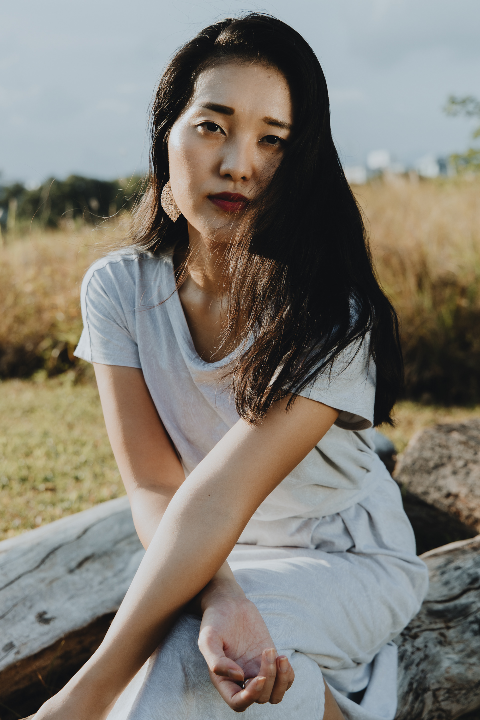 Vertical outdoor portrait of a woman in a light-colored dress, resting her arms on her knees, looking directly and intensely at the camera in sunset light.
