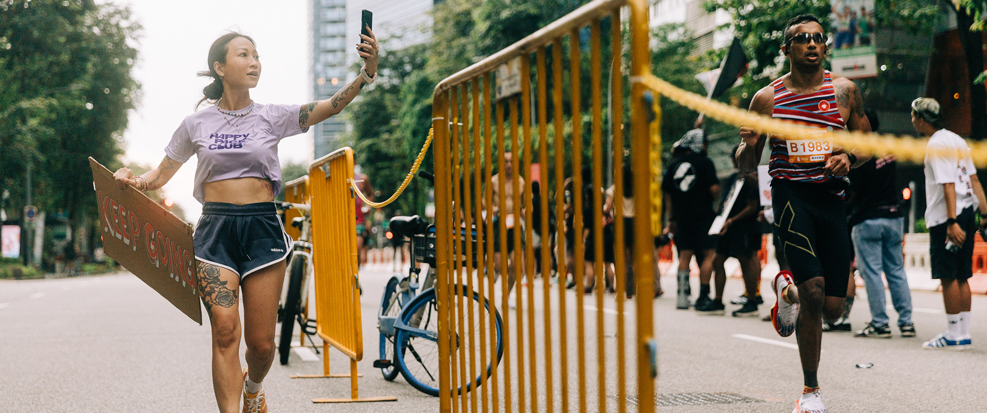 Cinematic wide shot of a focused marathon runner in Singapore passing a spectator recording a selfie against a yellow industrial barrier.