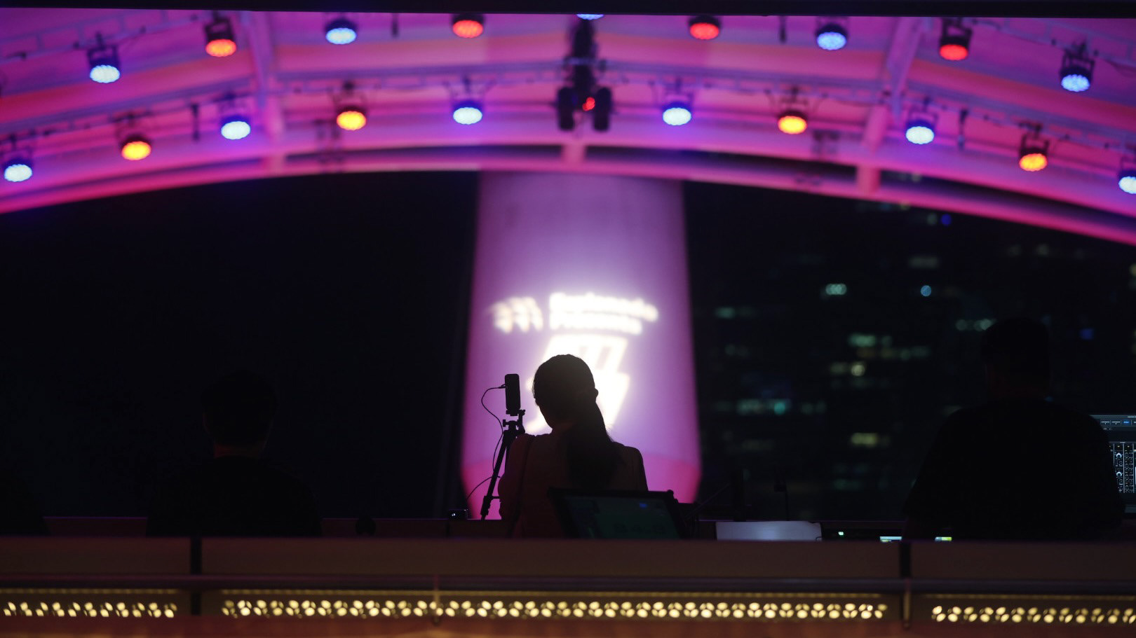 A cinematic 16:9 wide-angle extraction from a seated perspective at the Esplanade Outdoor Theatre. Dark silhouettes of a production crew and equipment occupy the foreground, framing a central illuminated pillar under vibrant purple and blue stage lighting. The shot emphasizes atmospheric bloom and the industrial grit of a live performance environment.